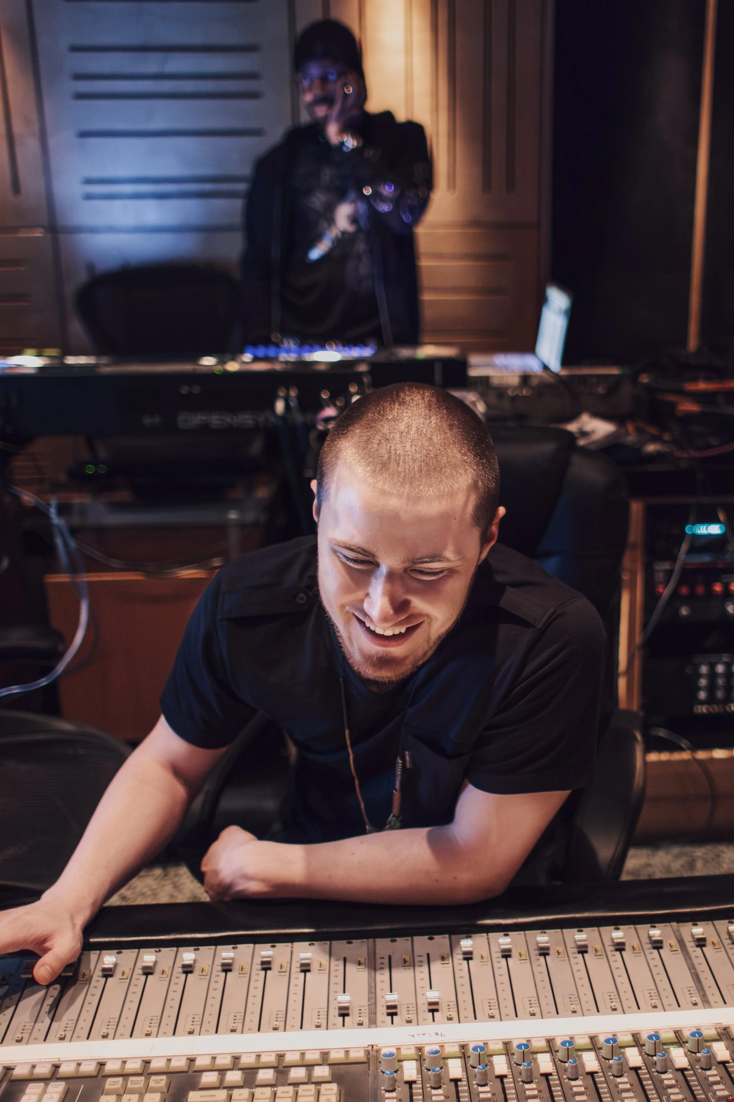 A man smiling in front of a sound mixing console, with a woman in the background near a computer monitor in a music studio.