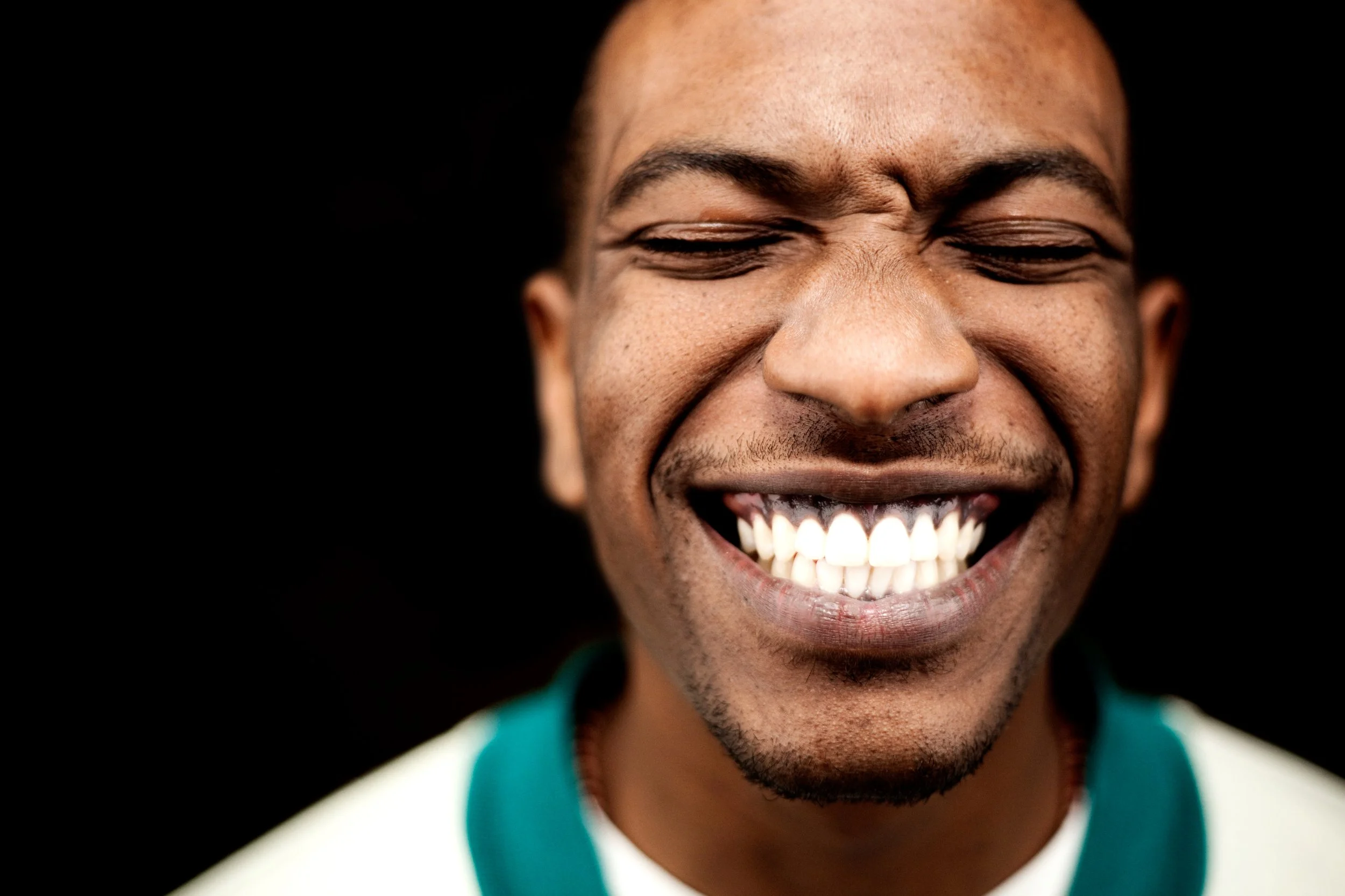 Close-up of a smiling man with closed eyes, showing his teeth, against a black background.