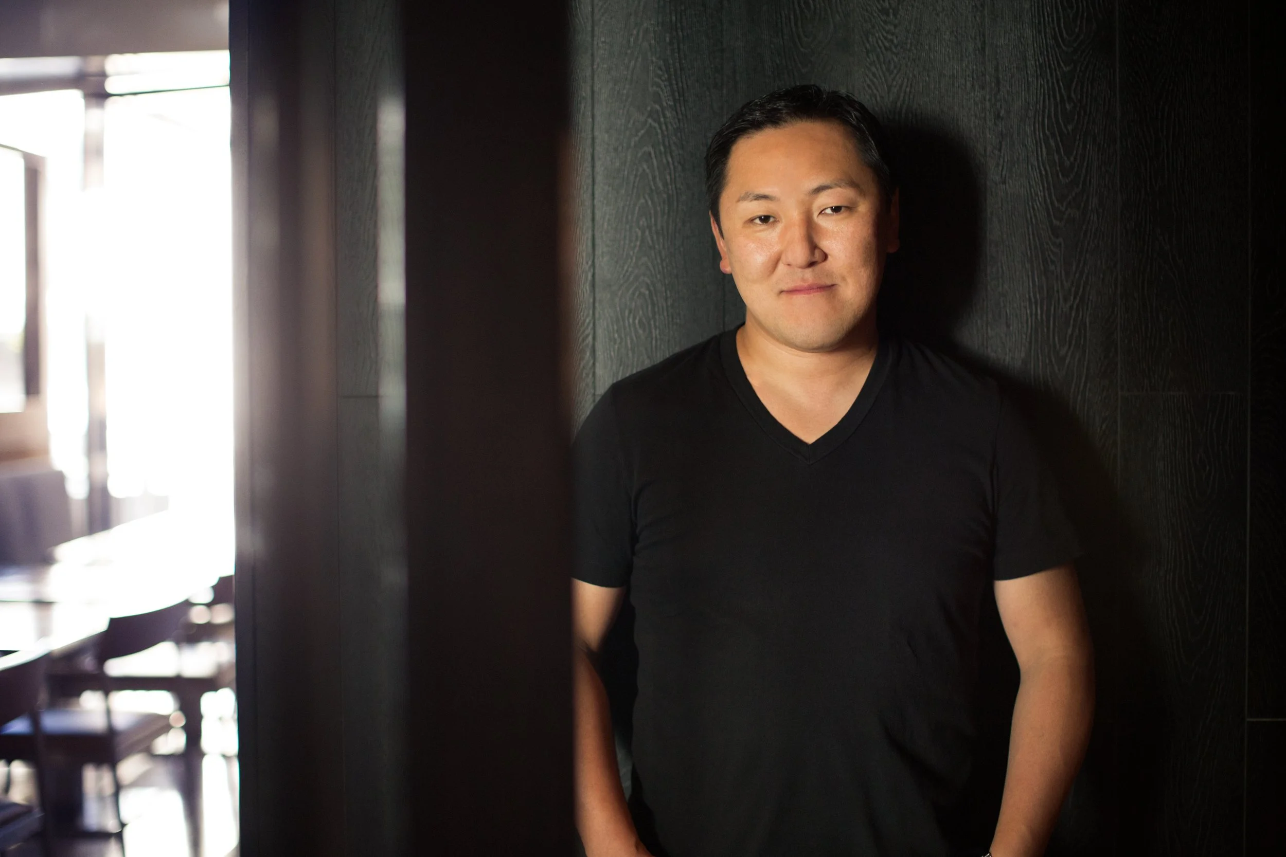 A man with short black hair and fair skin, wearing a black V-neck shirt, is standing against a dark wooden wall. He is slightly smiling and looking at the camera.