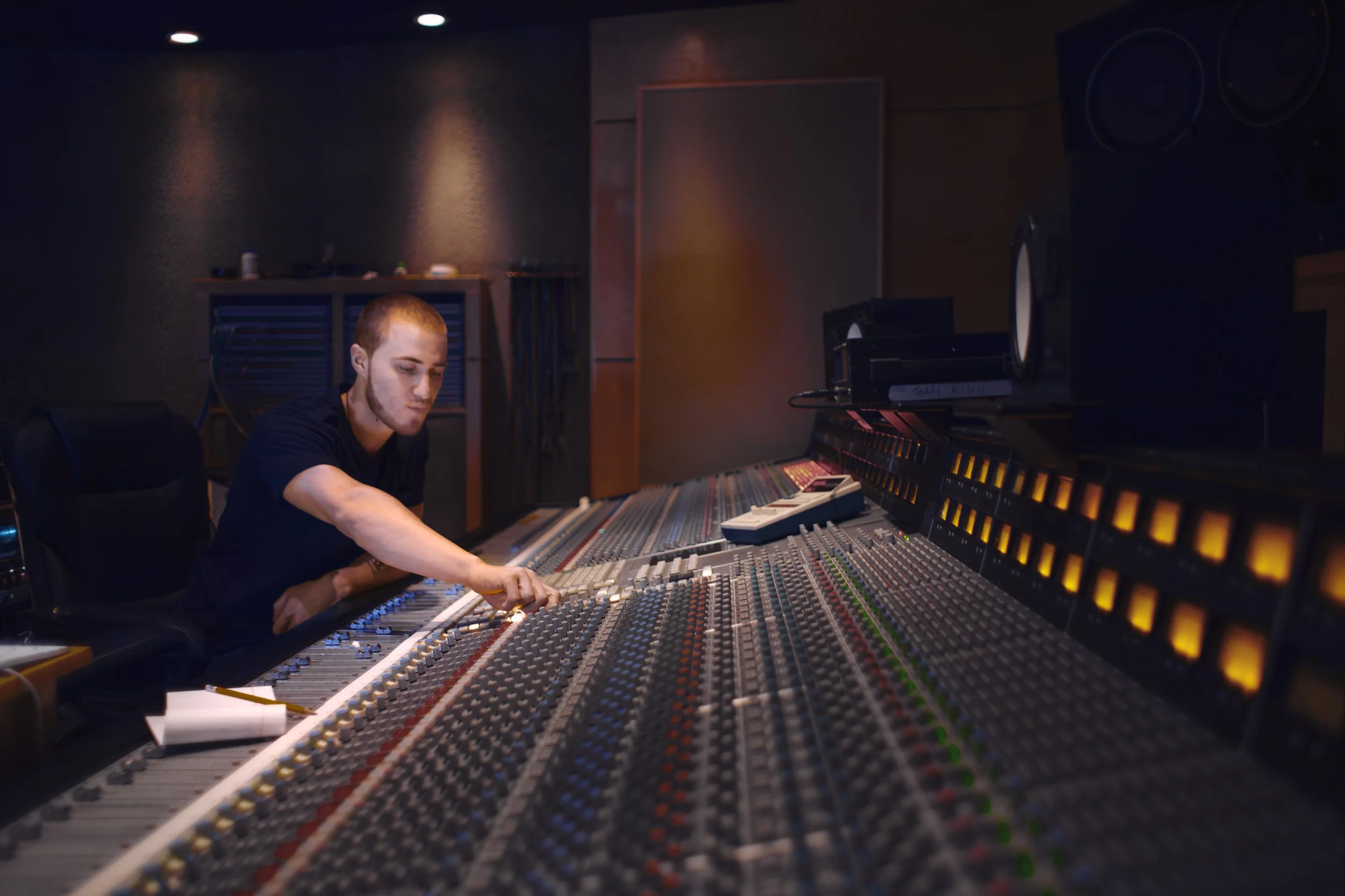 A person with a shaved head operating a large audio mixing console in a recording studio.