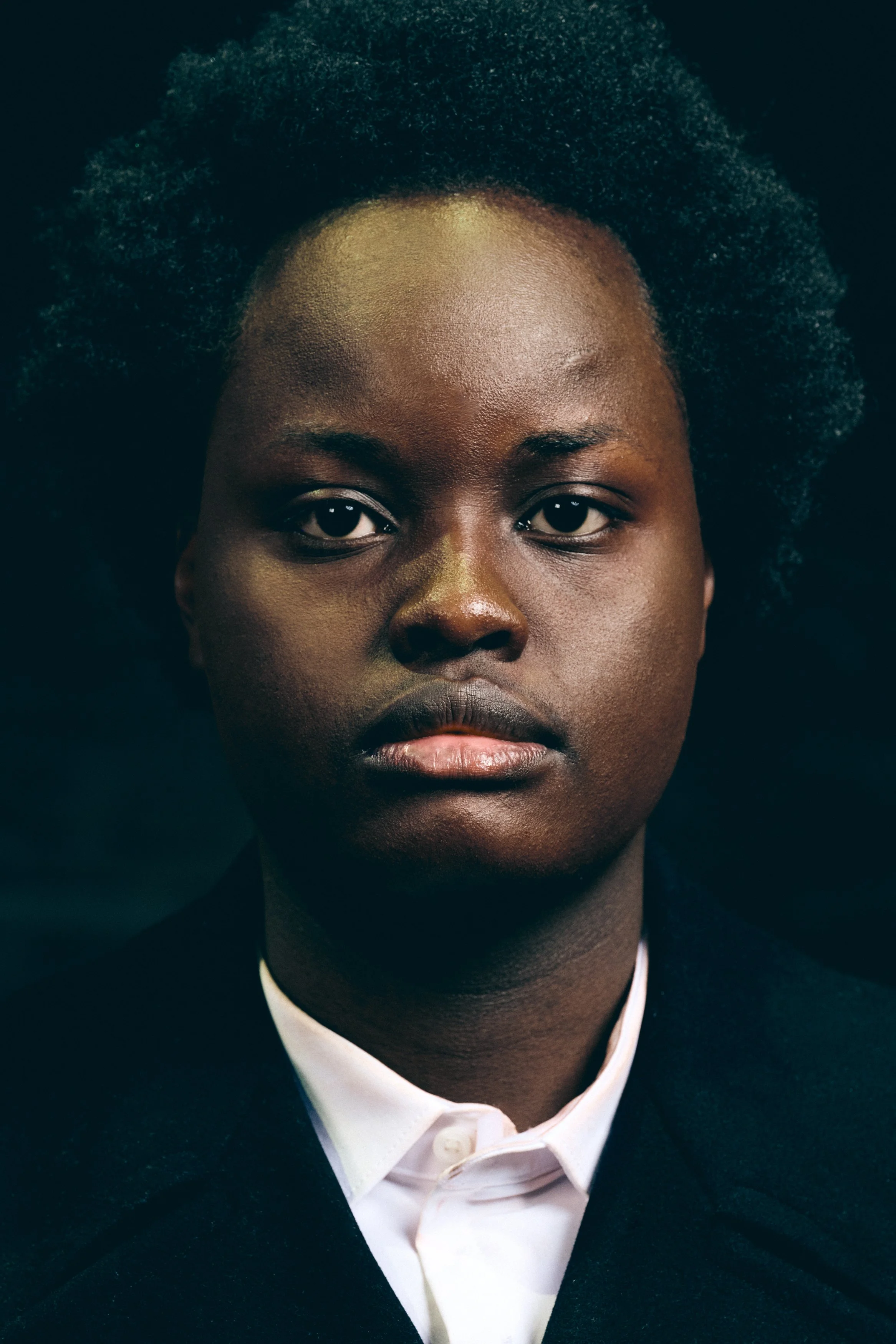 A close-up portrait of a Black woman with natural, textured hair, wearing a white shirt and dark blazer, against a dark background.