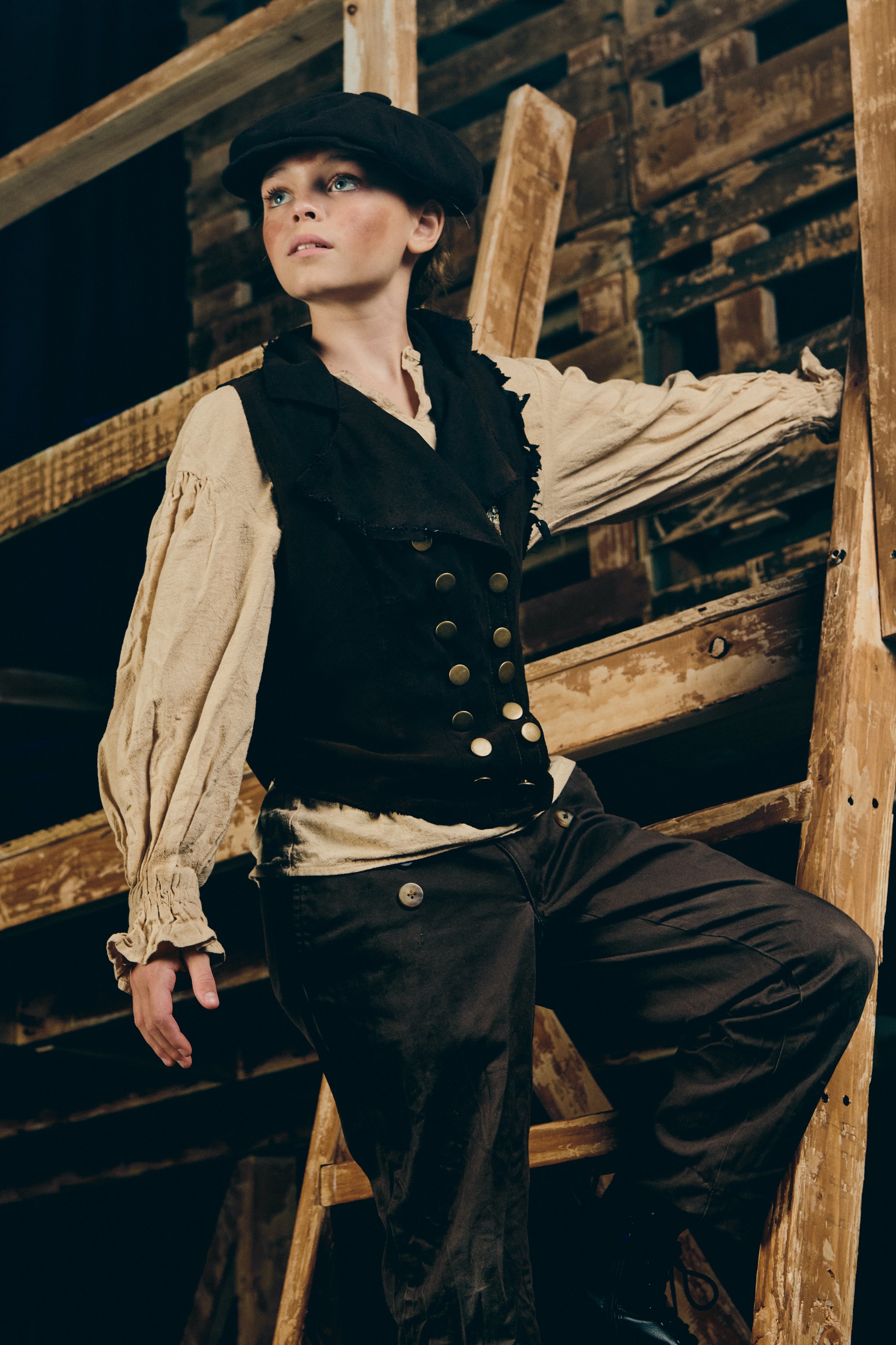 A woman leaning on a wooden staircase in a rustic setting, dressed in vintage-style clothing with a black beret, beige blouse with puffy sleeves, black vest with gold buttons, and black pants.