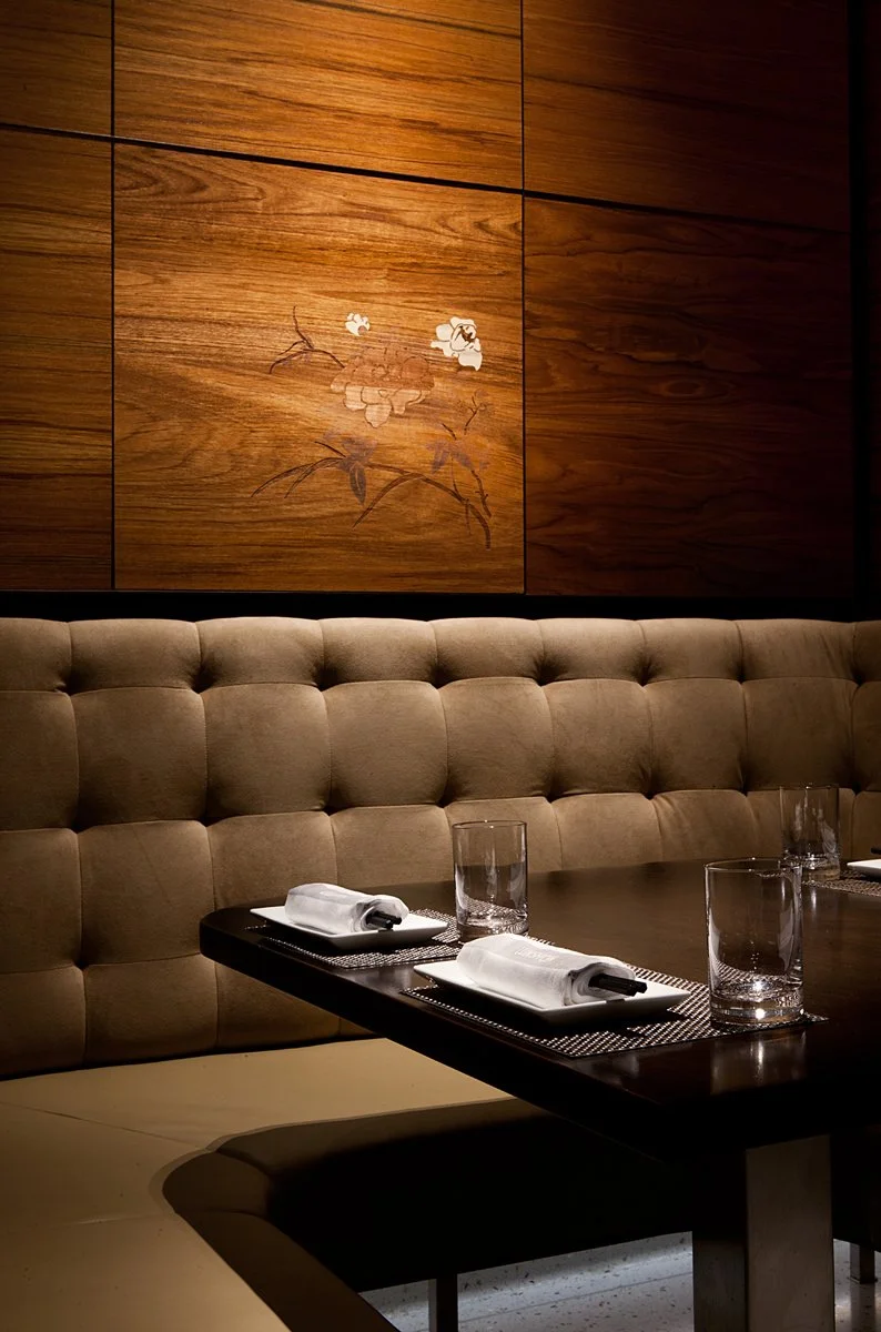 A restaurant corner with a tufted beige banquette, a dark wooden table set with white plates, rolled napkins, and empty glasses, against a wood-paneled wall with floral artwork.