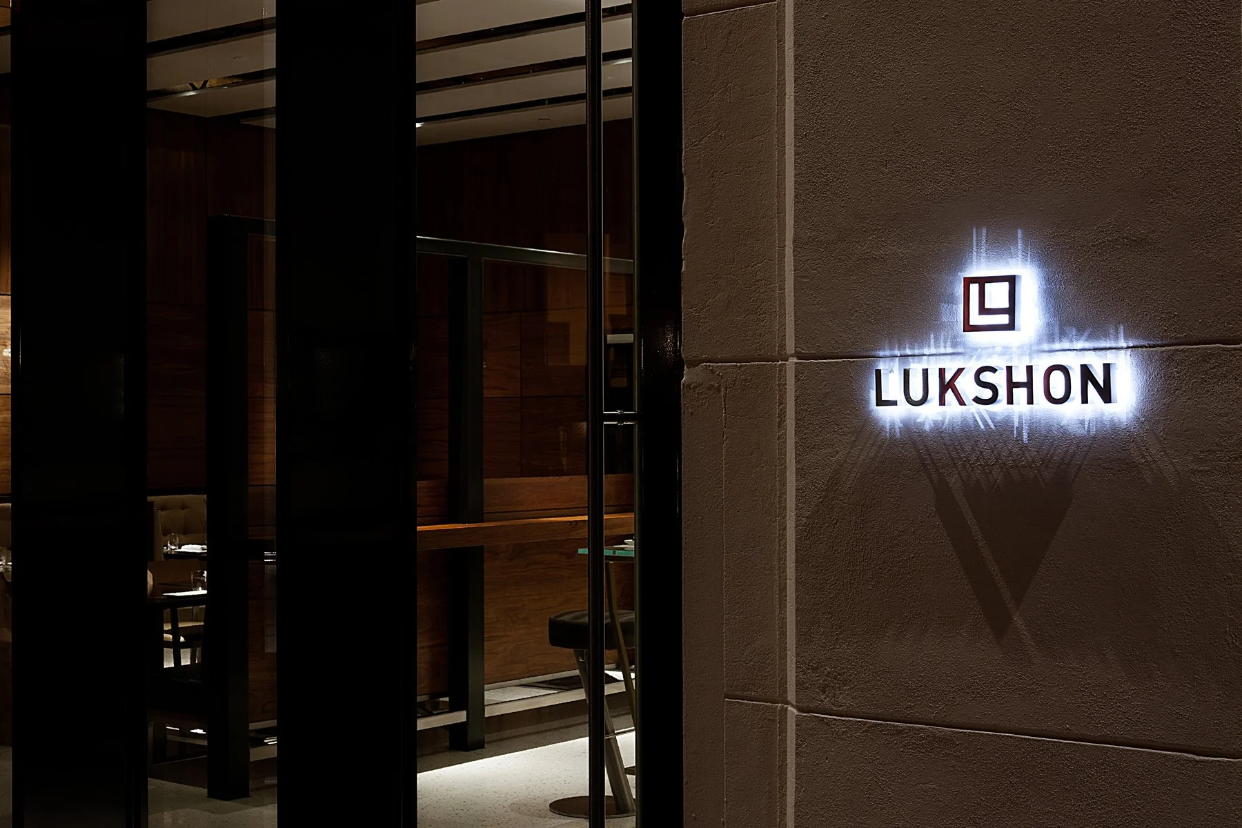 Nighttime view of a restaurant entrance with a lit sign reading 'LUKSHON' and a geometric logo on the wall, part of the interior visible through glass panels.