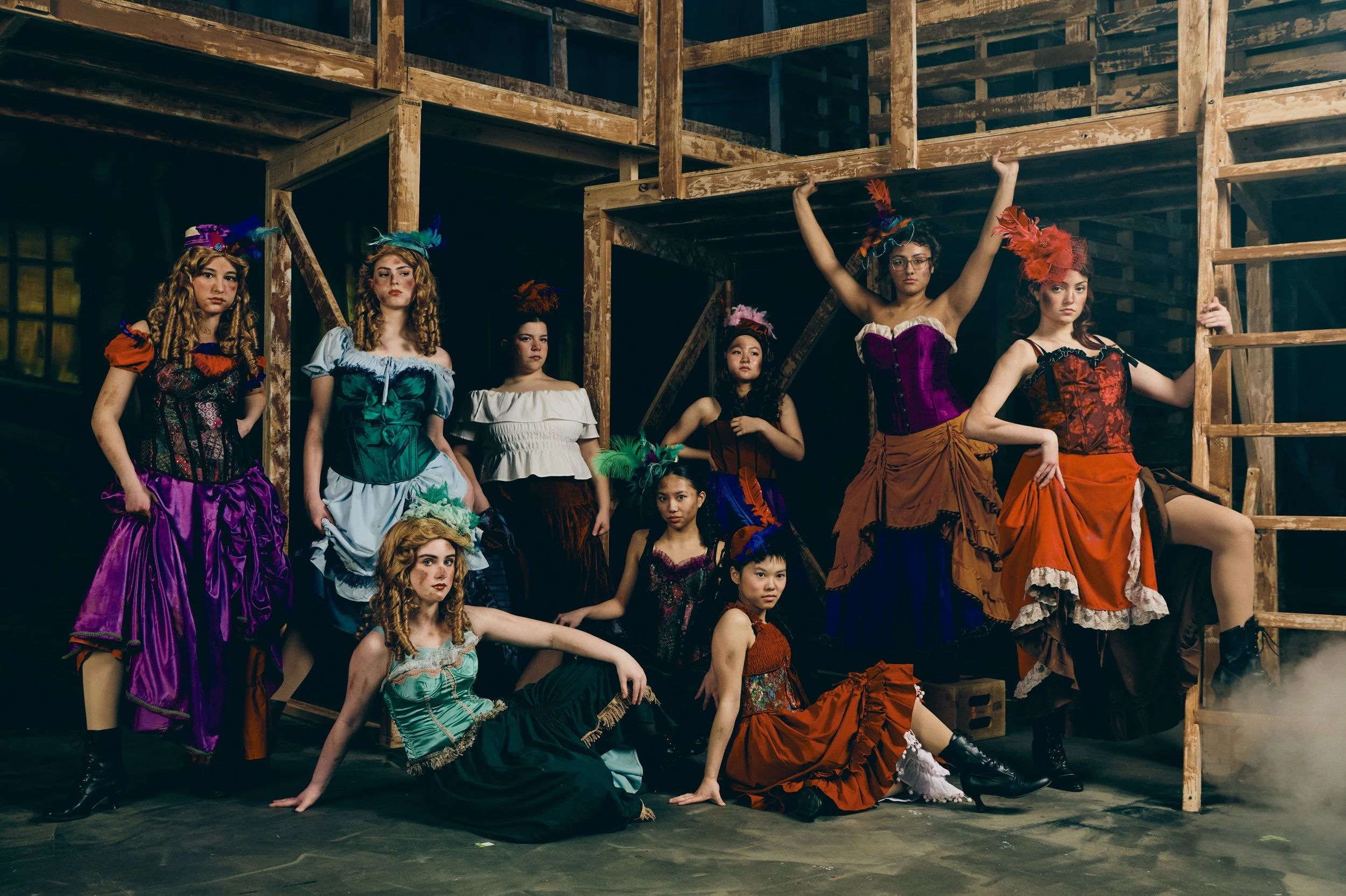 Group of women and girls dressed in colorful theatrical costumes, some with feathered hats, posing on a wooden stage framework in a theater or rehearsal space.