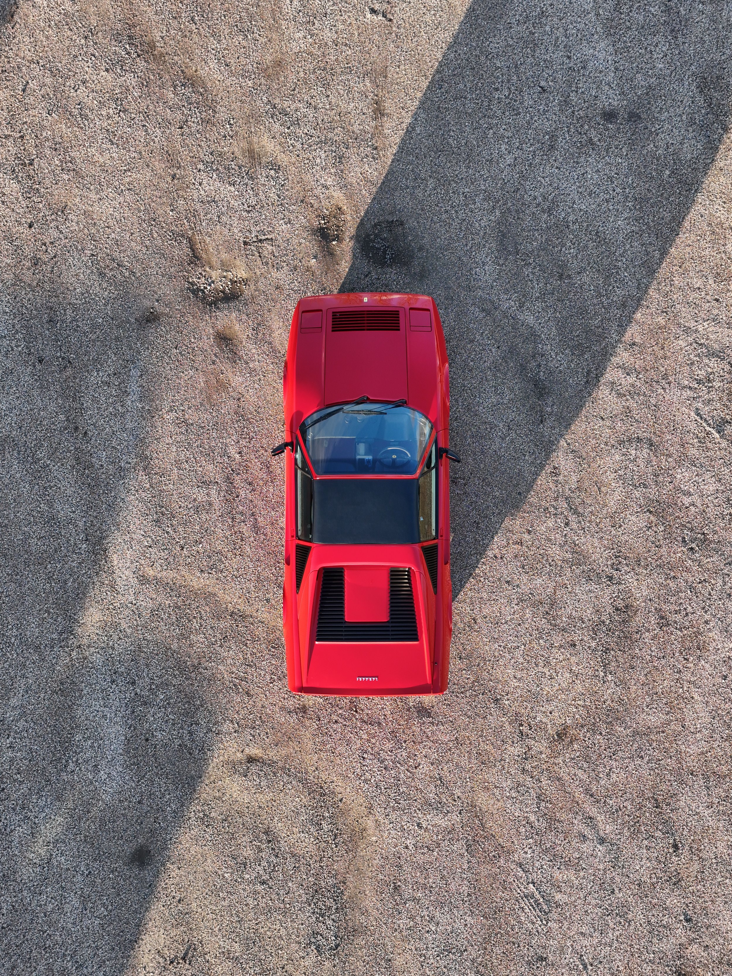 An overhead view of a red sports car on a gravel surface with a shadow cast to the right.