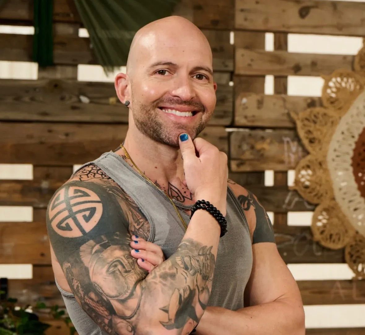 Smiling bald tattooed man in a gray tank top posing beside a person with blue nail polish