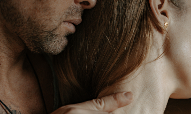 Close-up of a couple embracing, with a bearded man’s face near a woman’s neck and her hand resting on his shoulder.