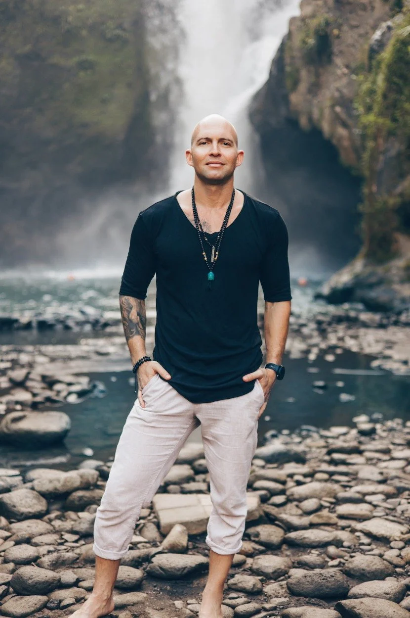 Bald man wearing a black shirt and necklaces standing in front of a waterfall between rocky cliffs