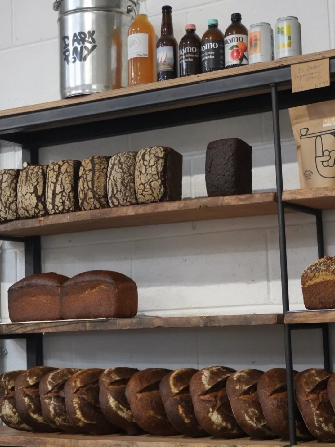 Wooden shelves displaying various types of bread, some with cracked crusts and others with smooth surfaces, against a white wall. Bottles of beverages and a metal container are on the top shelf.