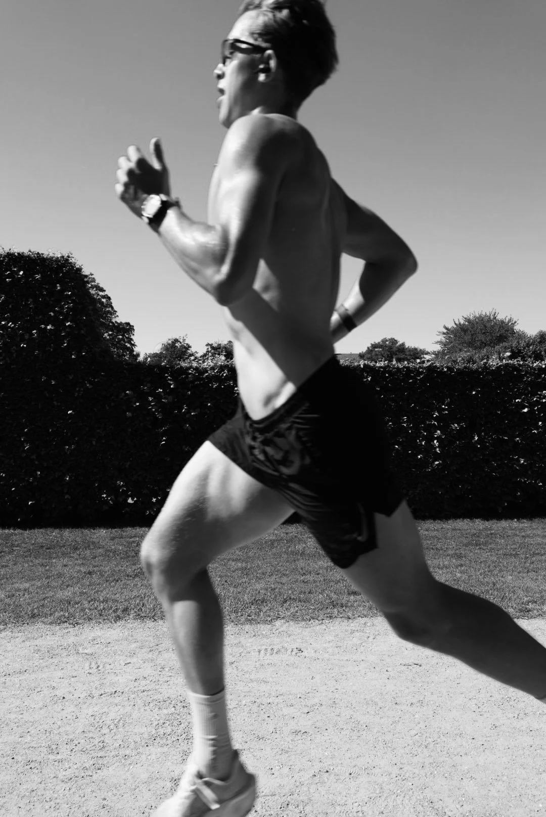 A man running outdoors on a dirt path, wearing glasses and athletic shorts, with trees and bushes in the background, captured in black and white.