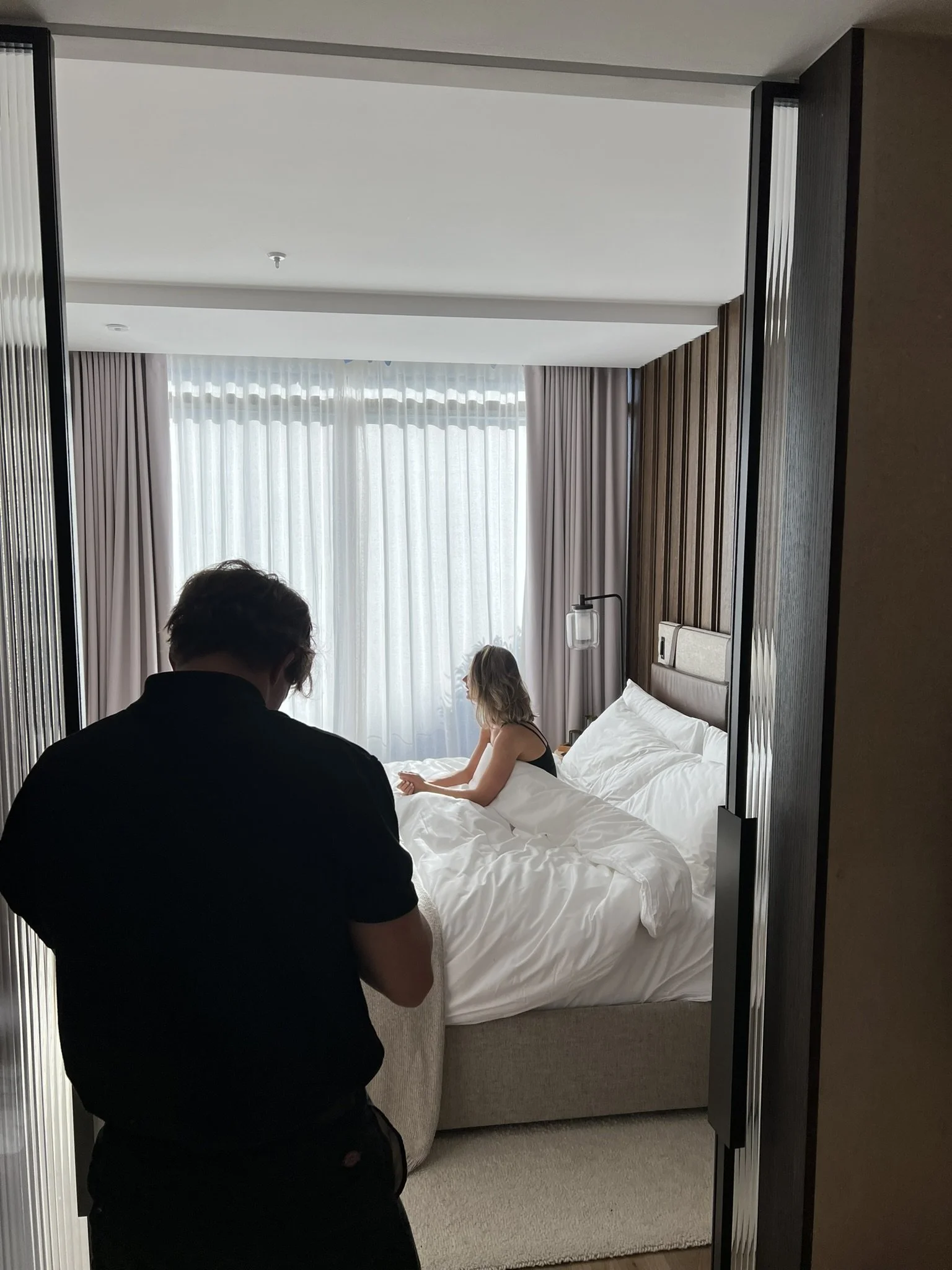 A person with short hair, wearing a black shirt, is standing in a doorway looking into a hotel room with a woman sitting on the bed. The room has large windows with sheer curtains and drapes, and wooden wall paneling.