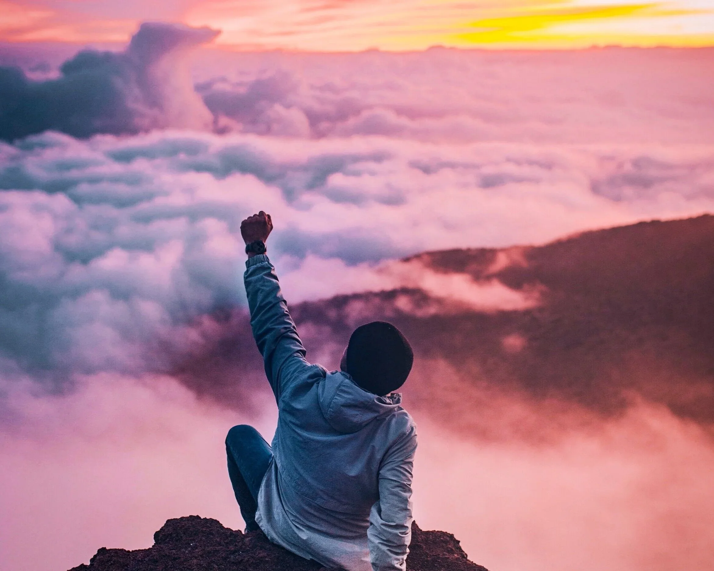 Person sitting on a rock at the top of a mountain, raising their fist in front of a colorful sky filled with clouds during sunset.