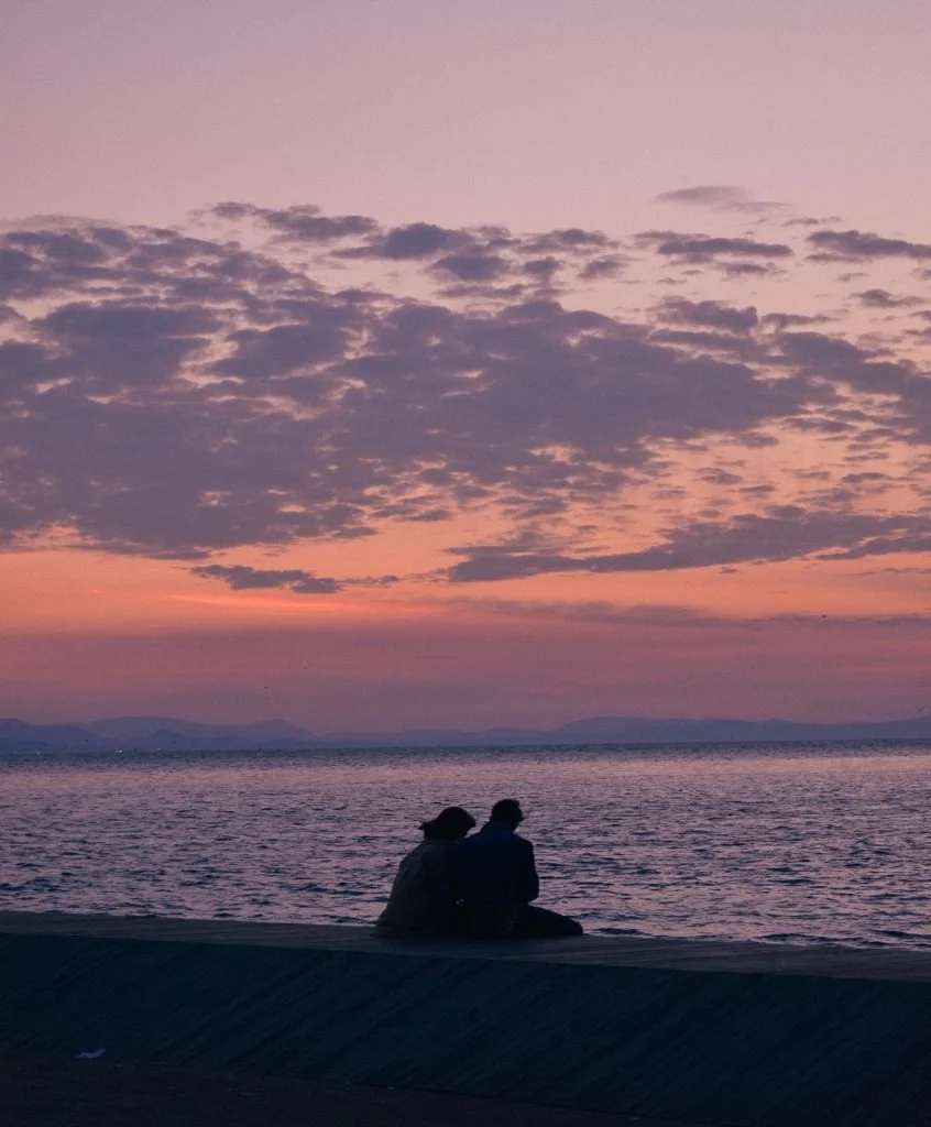 Two people sitting on a concrete ledge by the water, watching a sunset with pink and purple clouds over a calm lake or sea.