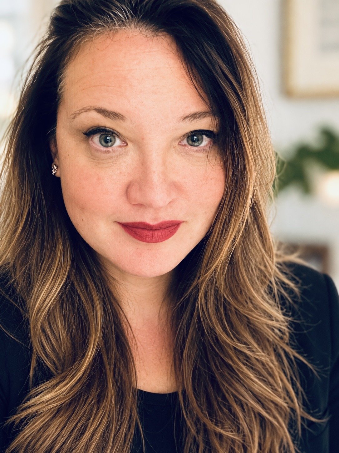 Close-up portrait of a woman with long, wavy brown hair, freckles, blue eyes, red lipstick, and wearing a black top and floral earrings.