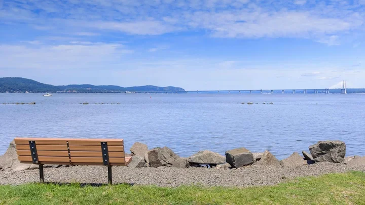 A wooden bench facing a calm body of water, with rocks lining the shore and a bridge visible in the distance under a partly cloudy sky.