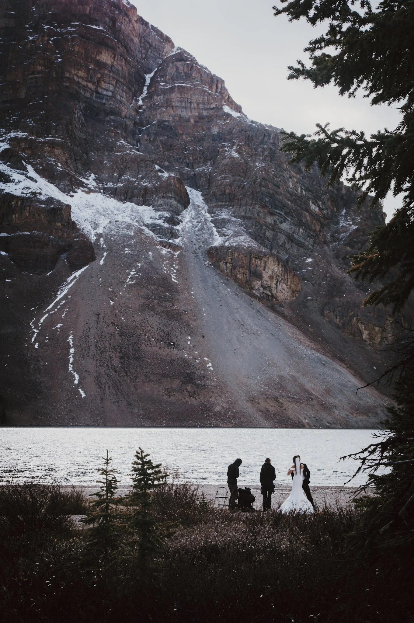 A group of four people, including a wedding bride in a white dress, near a lake with a mountain in the background, taken during dusk or early evening.