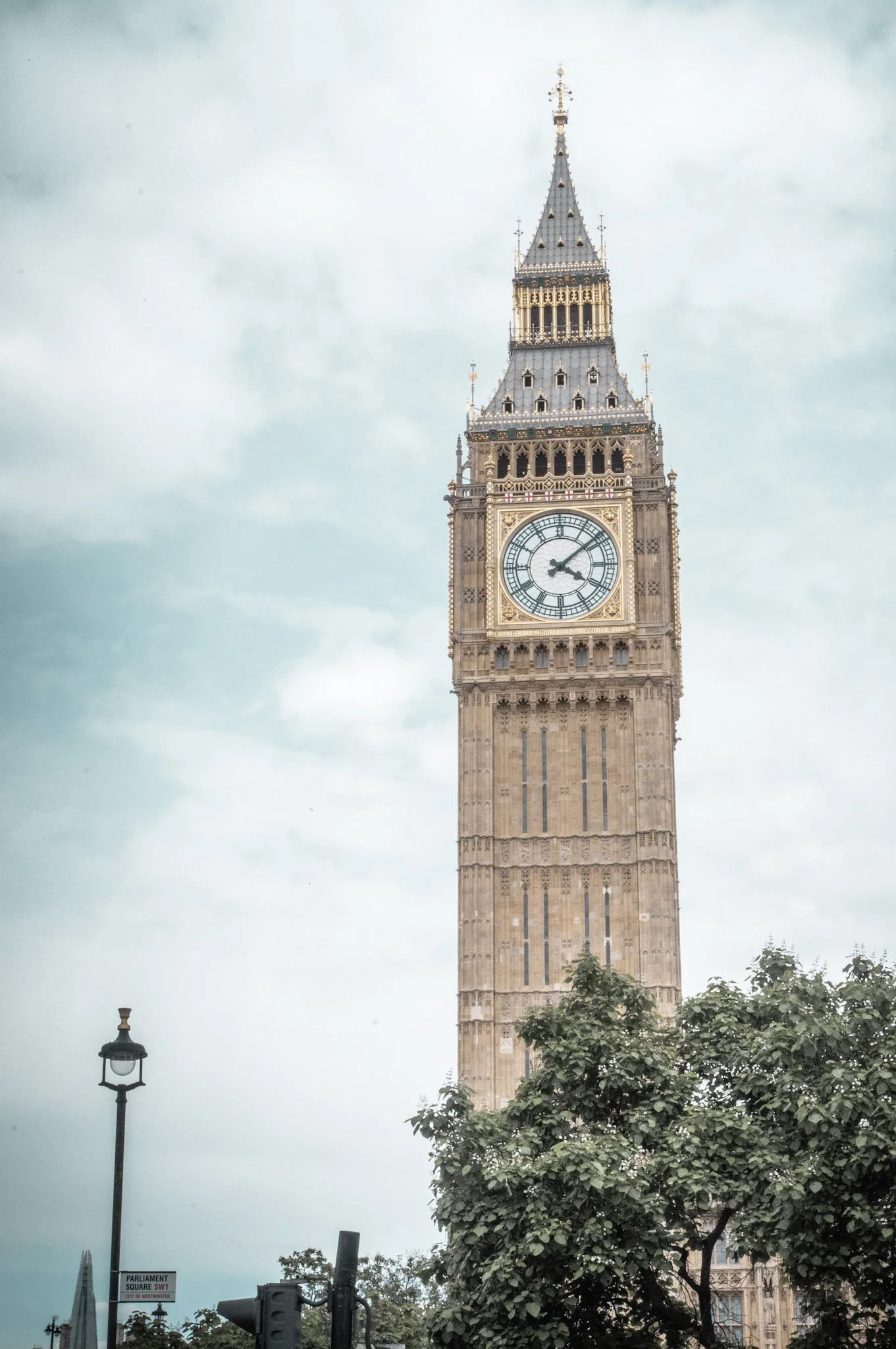 A view of Big Ben clock tower in London, showing the time as approximately 3:10, with cloudy sky and tree branches in the foreground.