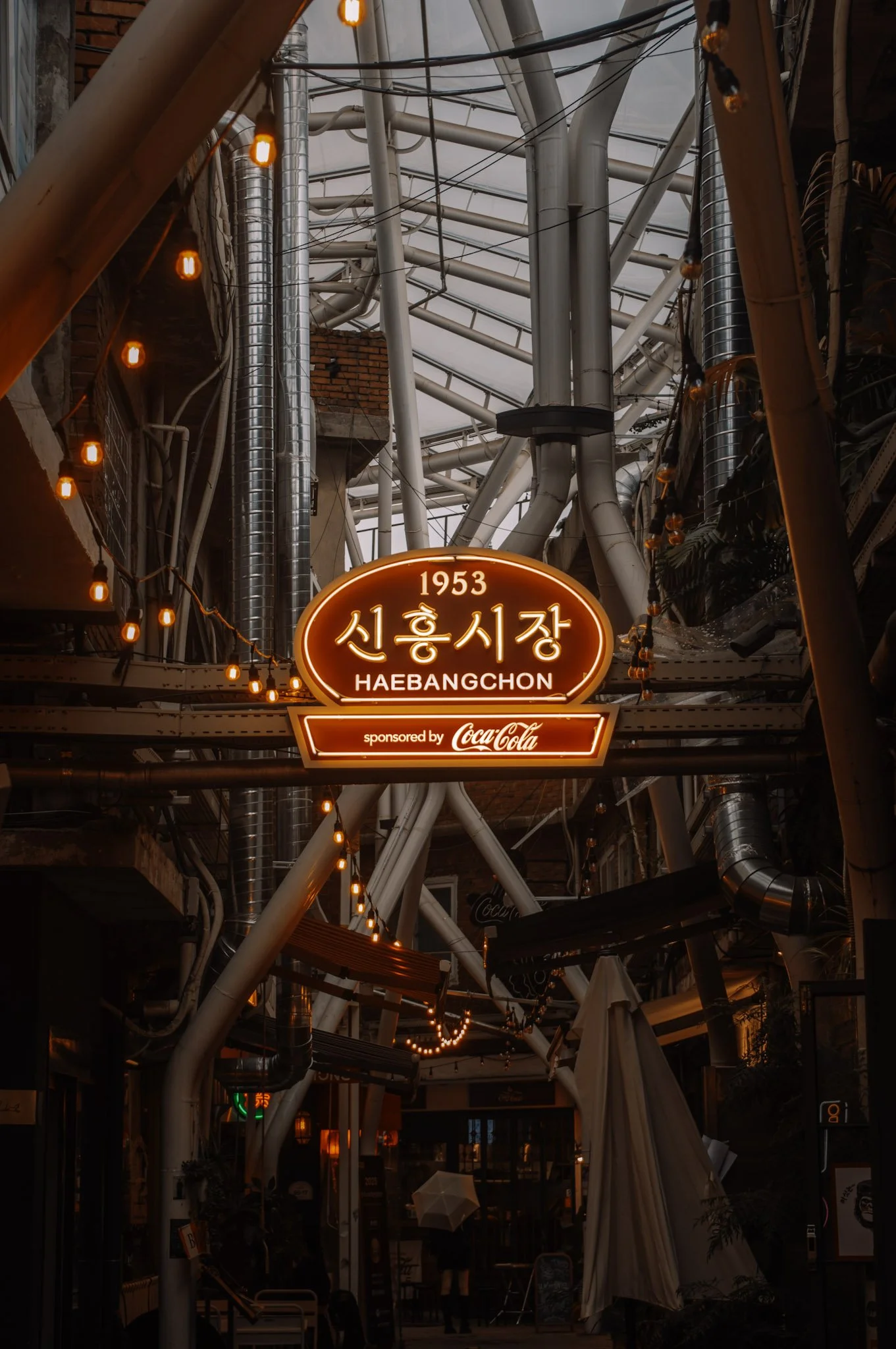 A covered outdoor market with hanging string lights, metallic ventilation pipes, and a sign in Korean and English reading '1953 Sinheung Market Haebangchon,' sponsored by Coca-Cola.