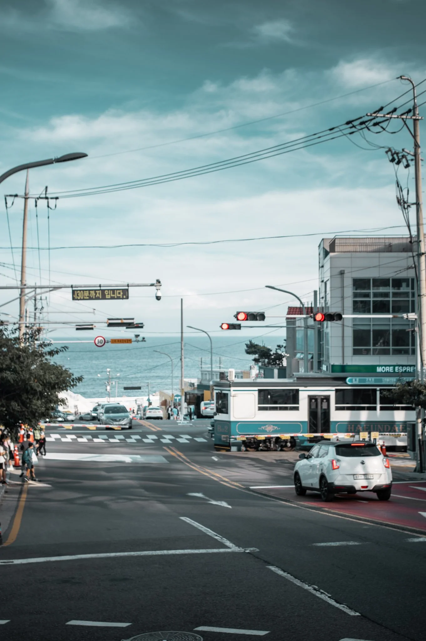 A busy city street scene with cars, pedestrians, traffic lights, and overhead power lines with mountains and sky in the background.