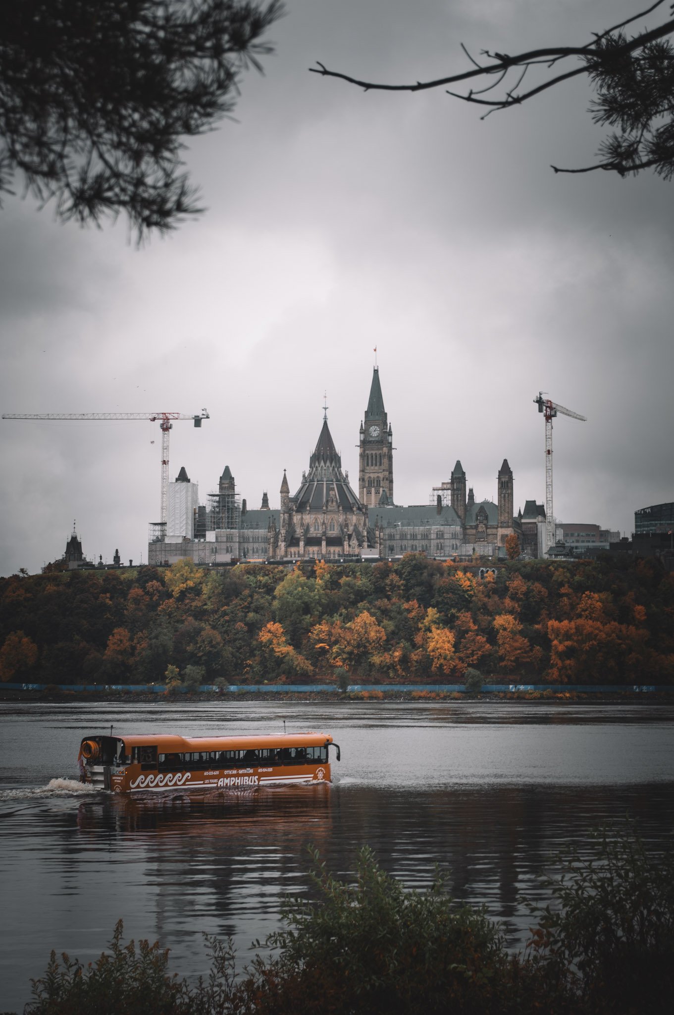 A cityscape featuring a large historic building with clock towers, possibly Parliament, under a cloudy sky. Construction cranes are visible on either side, and a orange tour boat labeled 'Amphibus' is moving across a river in the foreground. Autumn t