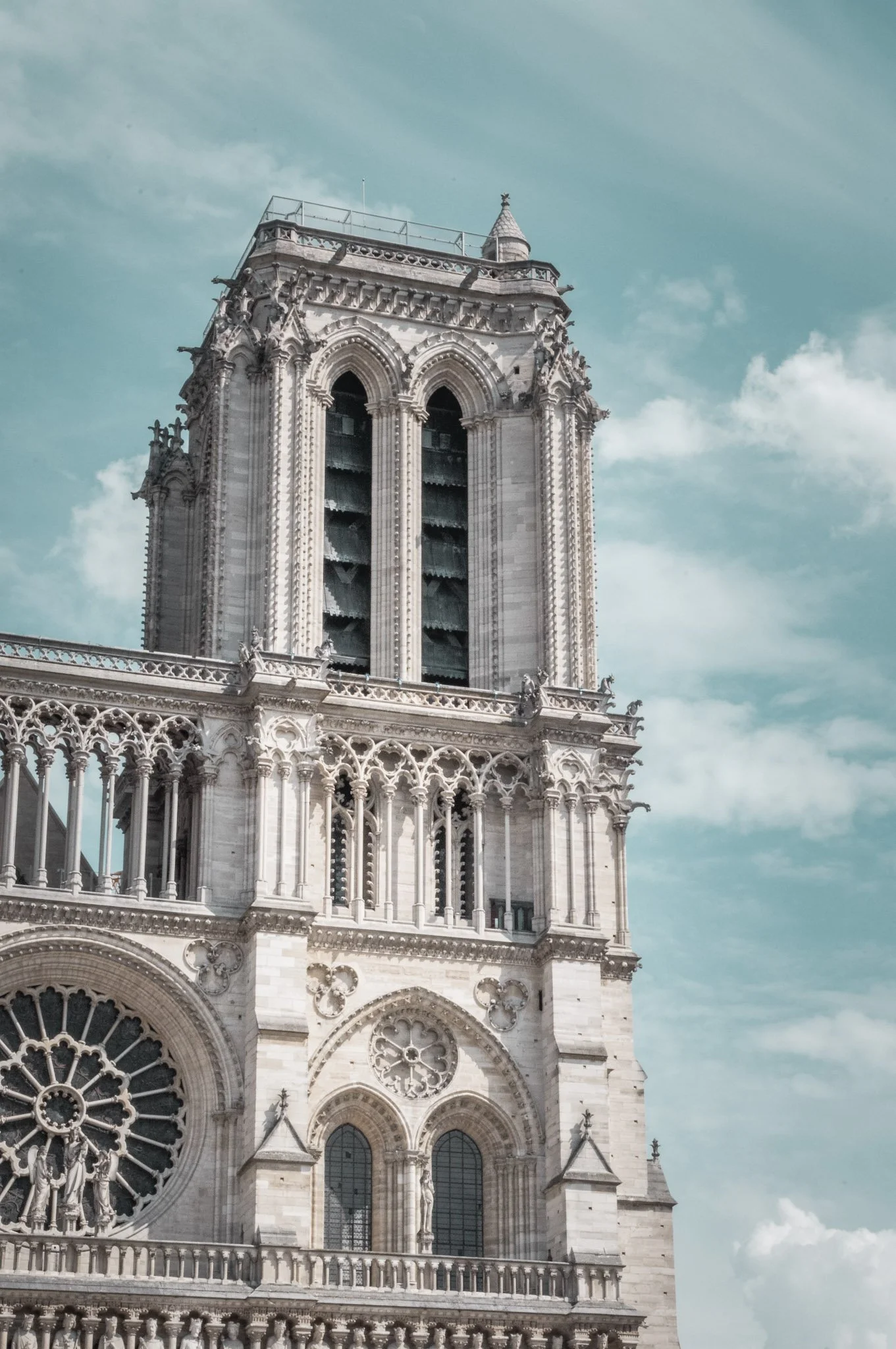 Close-up of the front of the Notre-Dame Cathedral showing its intricate Gothic architecture, stained glass windows, and spire against a partly cloudy sky.