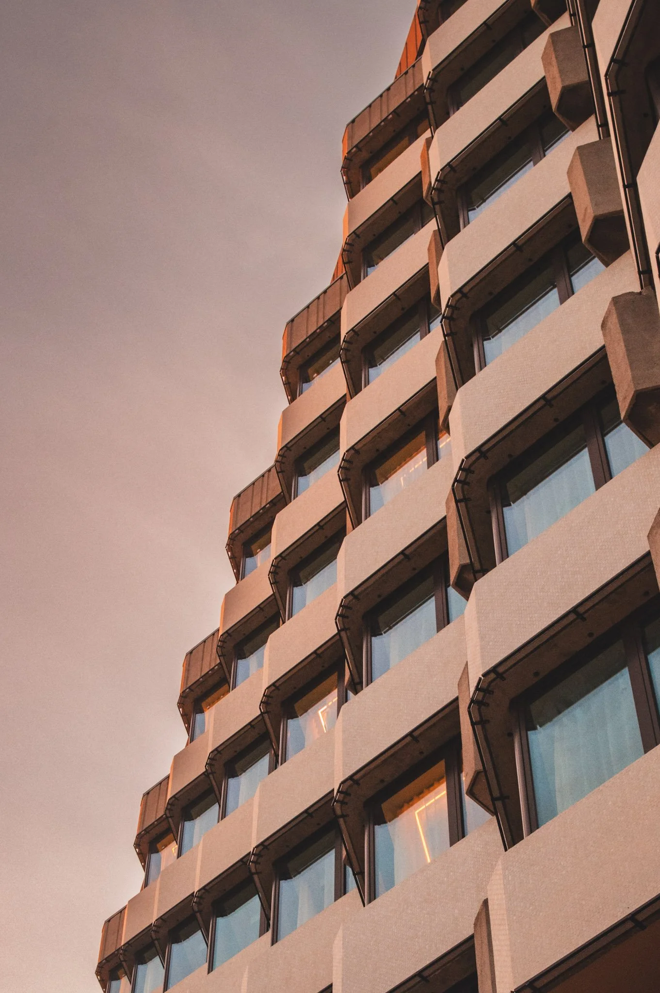 A modern multi-story residential building with curved balconies and glass windows, illuminated by warm lighting at sunset or dusk.