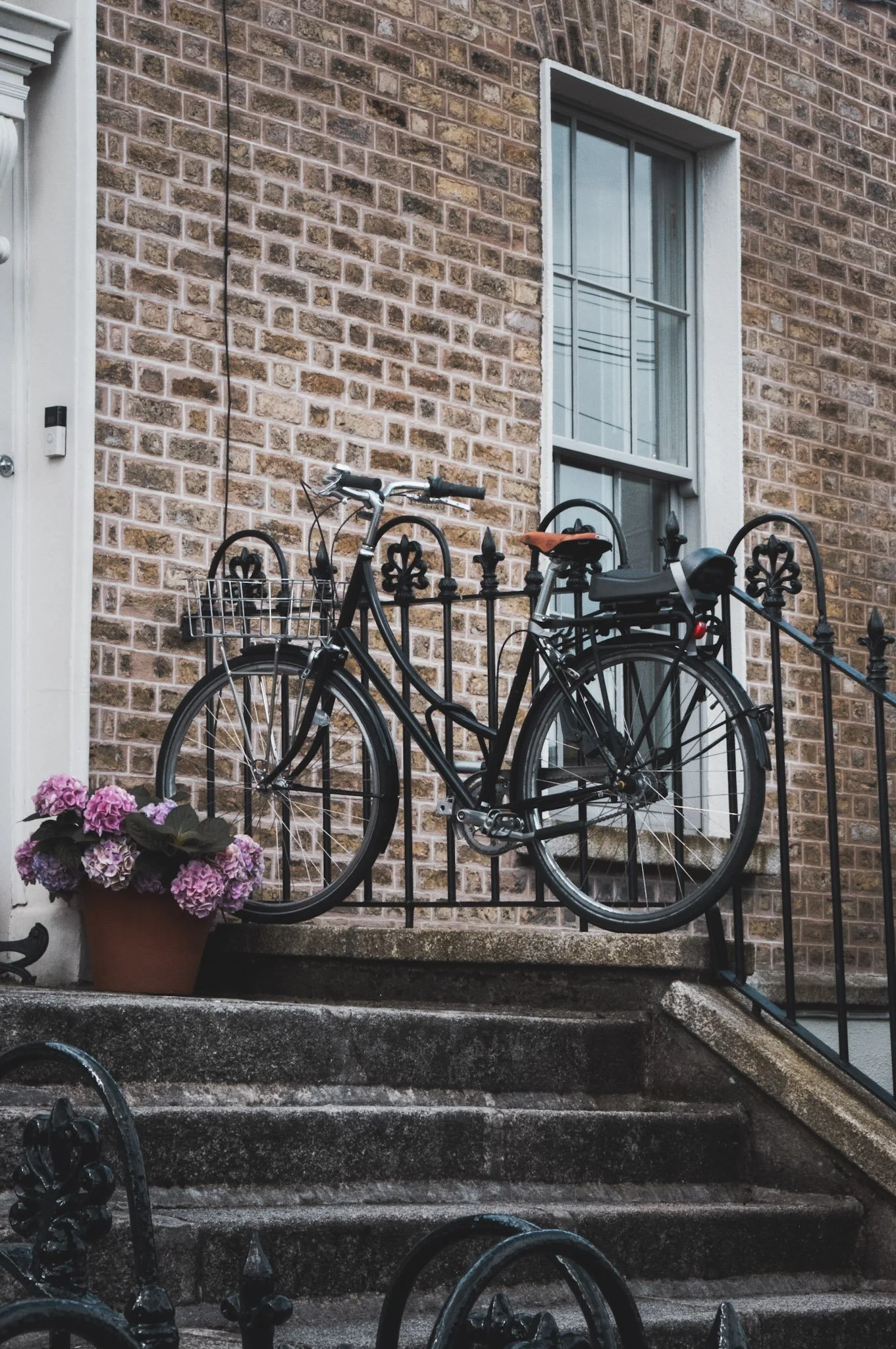 A black bicycle with a front basket is parked in front of a brick house with a window. Pink hydrangea flowers are in a corner pot near the bicycle. The house has a white doorframe with a doorbell on the side, and black iron fencing surrounds the smal