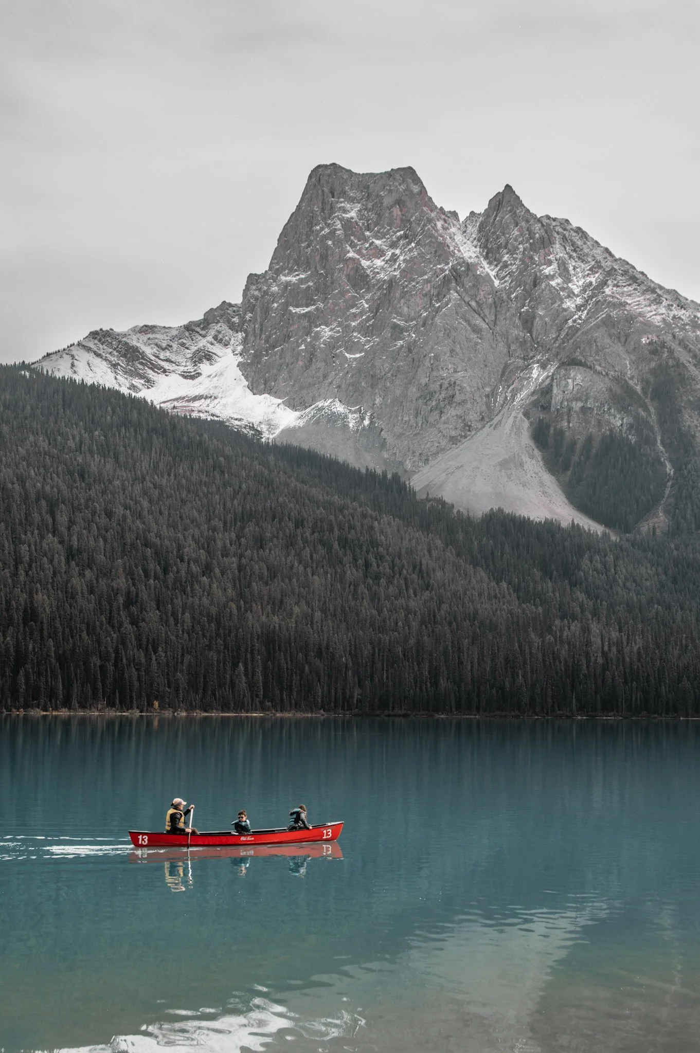 A small red boat with three people on a calm turquoise lake, with a backdrop of dense evergreen forest and large rugged mountain peaks with patches of snow, under a cloudy sky.