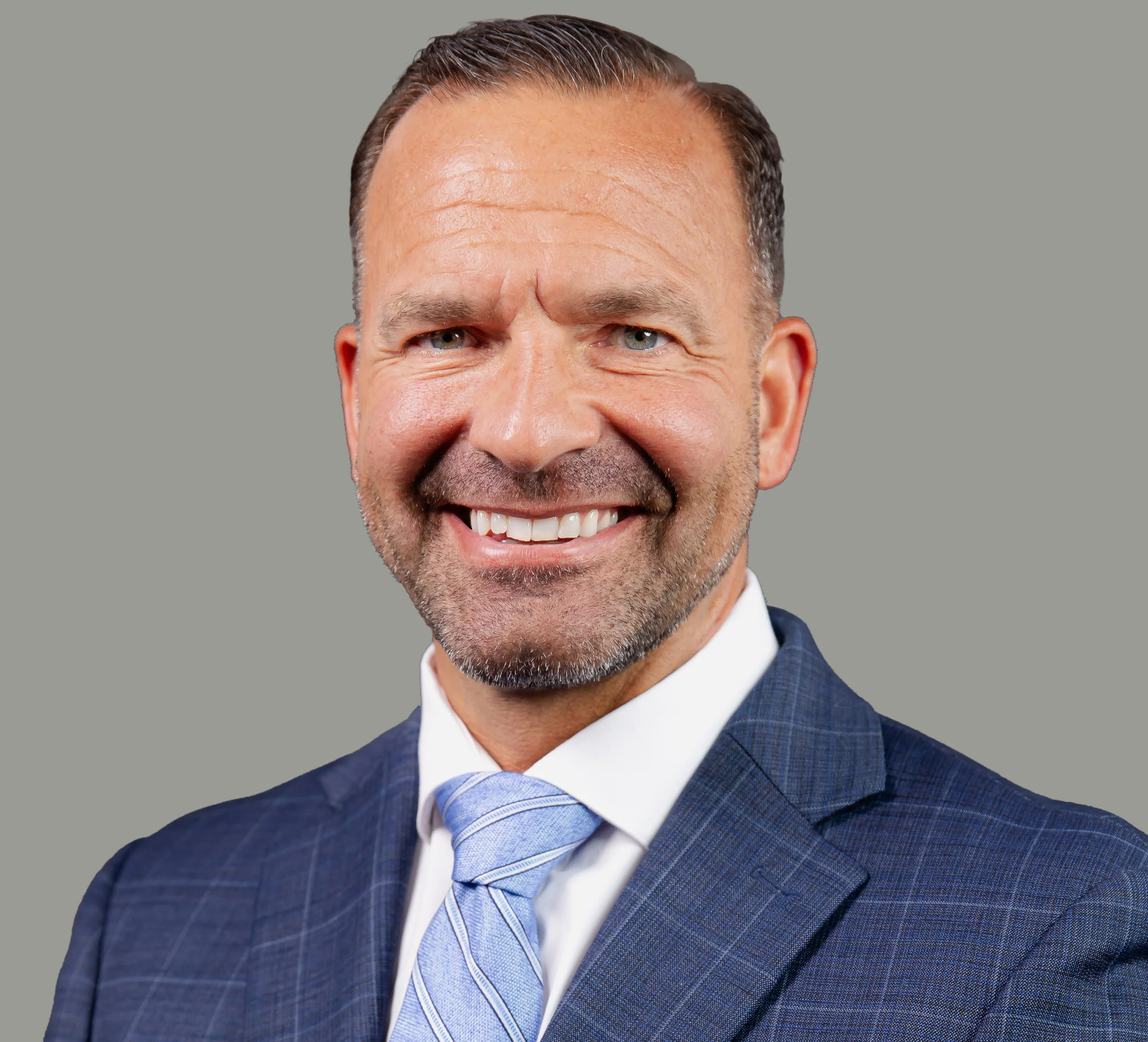 Close-up of a smiling man in a blue checkered suit with a light blue tie, against a plain gray background.