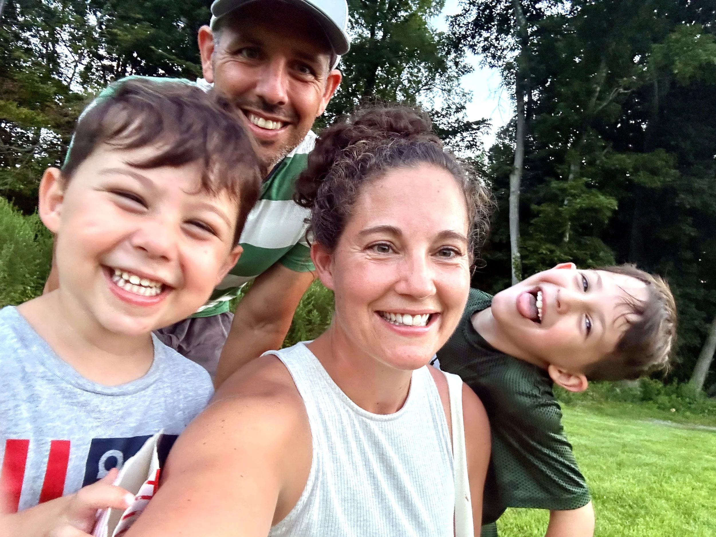 Smiling family of four taking a selfie outdoors with trees in the background.