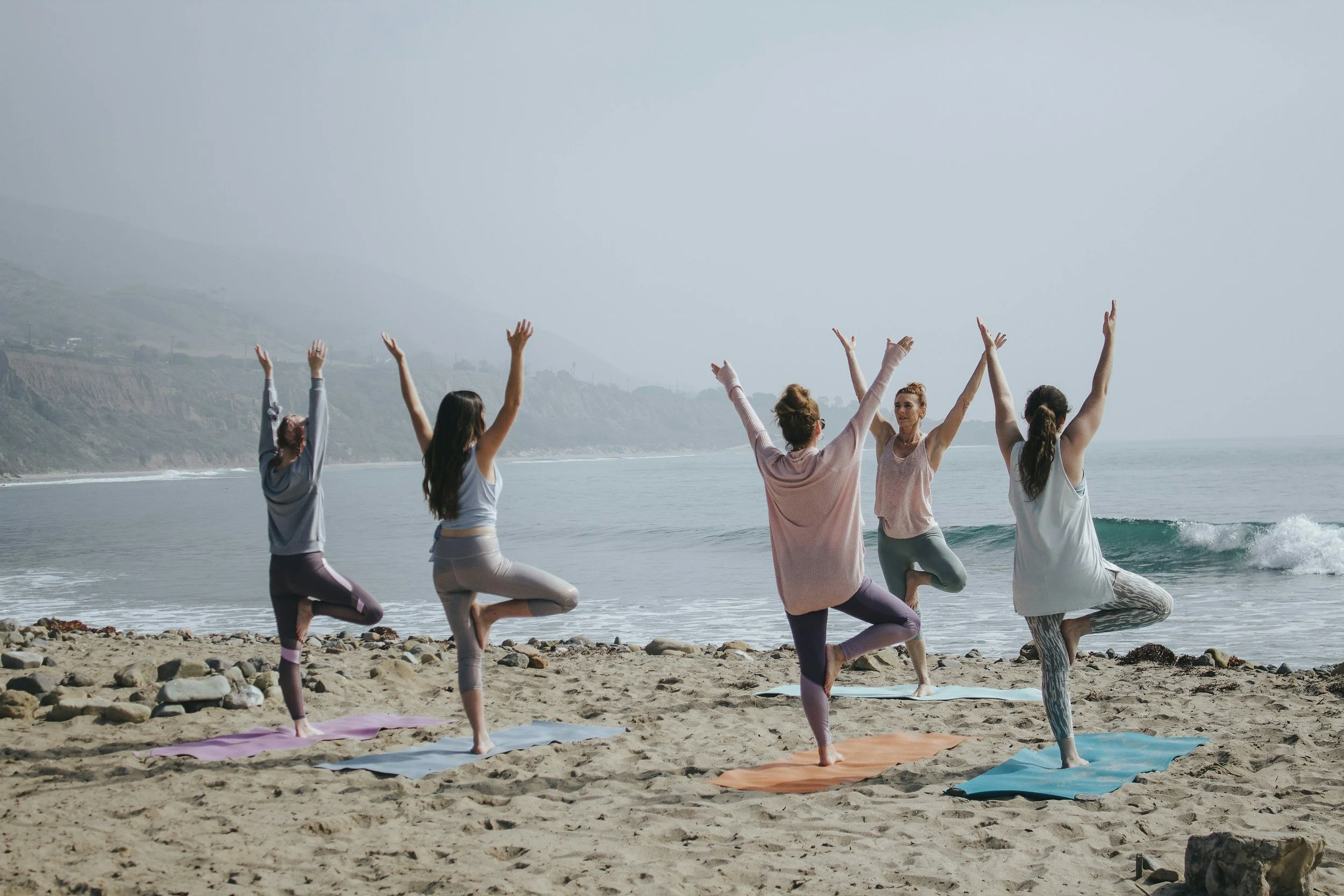 Group of diverse women practicing yoga on the beach with ocean and hills in the background.