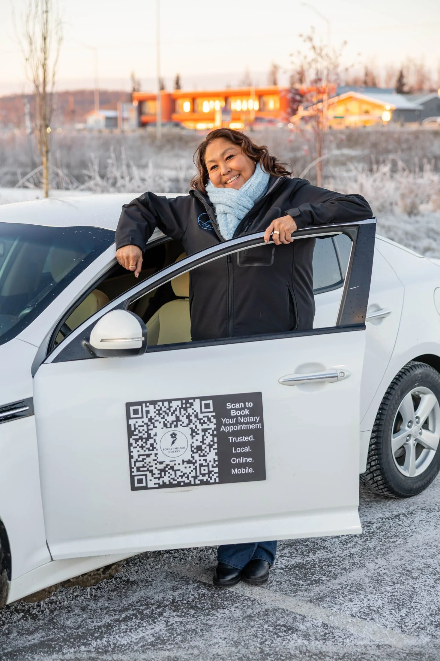 A woman smiling and leaning out of a white car's window during winter. She is wearing a black jacket, blue scarf, and has short curly hair. The car has a QR code and text on the side offering notary services, with a snowy outdoor background and buildings in the distance.