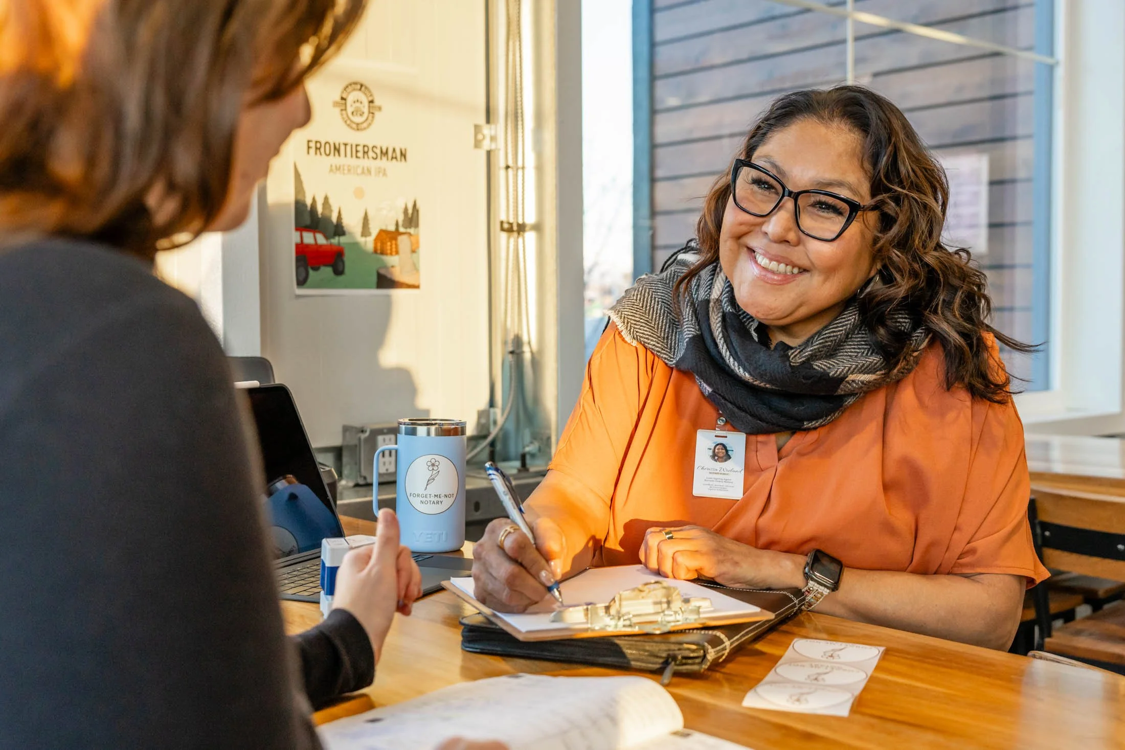 A woman with glasses and a scarf smiling as she talks to another person at a table with documents and a pen.