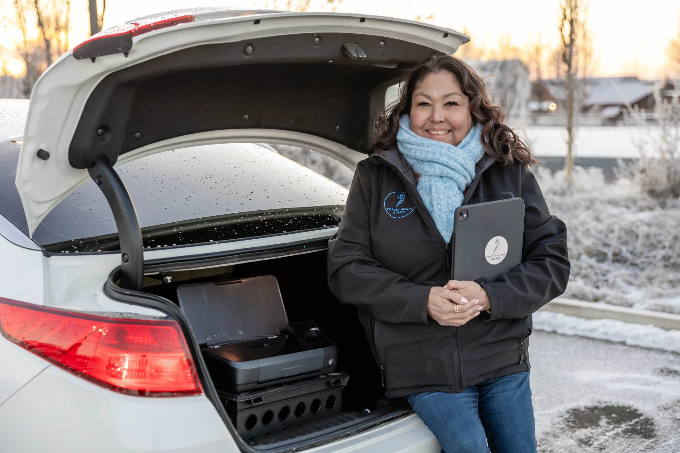 A woman with curly brown hair smiling, standing outdoors next to an open car trunk, holding a tablet or notebook with a logo that says 'Forget Me Not Notary.' She is wearing a black jacket, a blue scarf, and jeans. The scene appears to be during sunset or sunrise in a winter setting with snow on the ground and trees in the background.