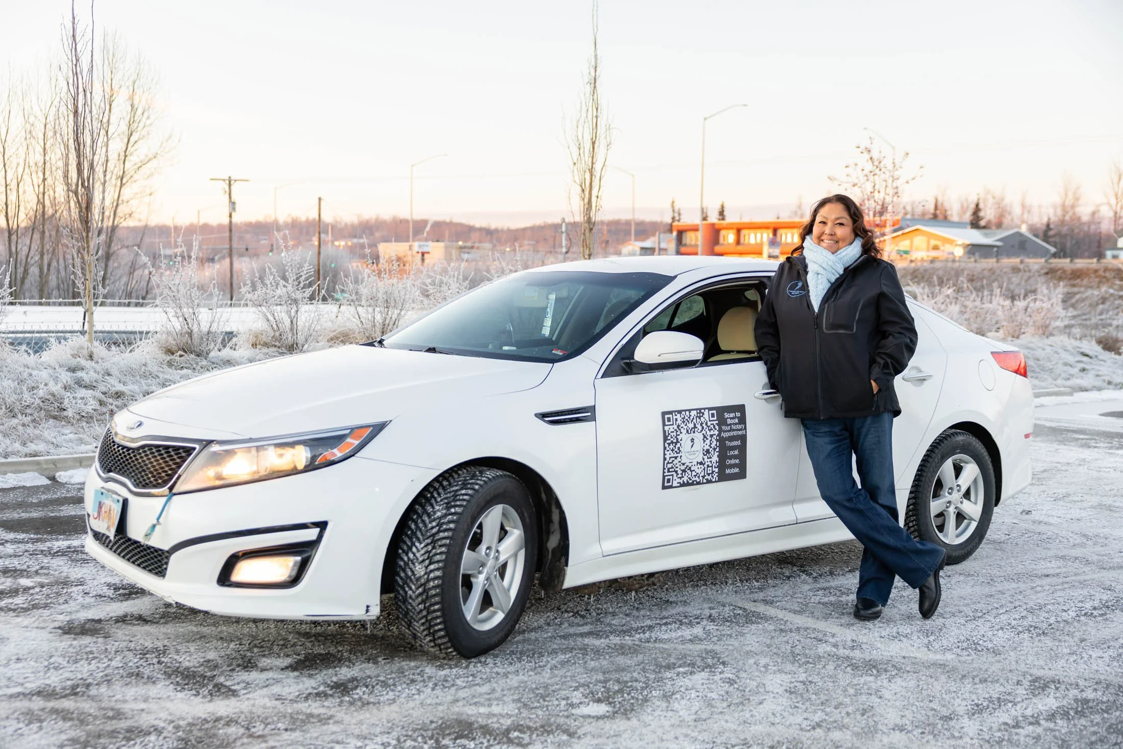 A woman standing next to a white car in a snowy parking lot during winter, smiling and wearing a black jacket and a light blue scarf.