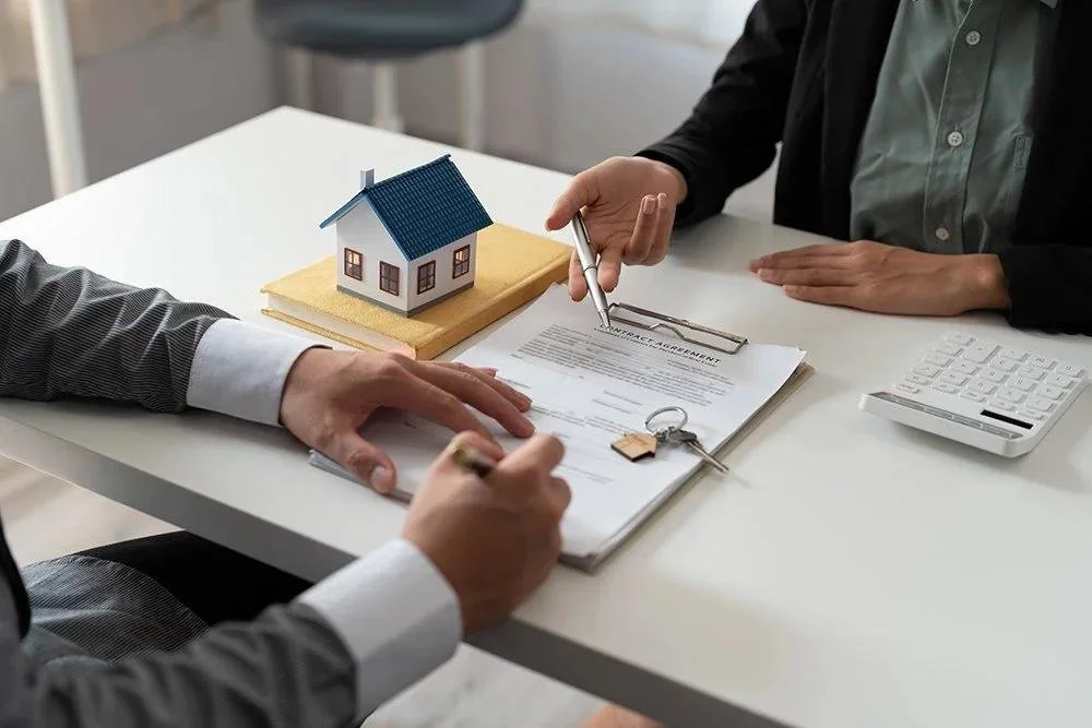 Two people signing a contract at a desk with a miniature house model on it, suggesting a real estate transaction.