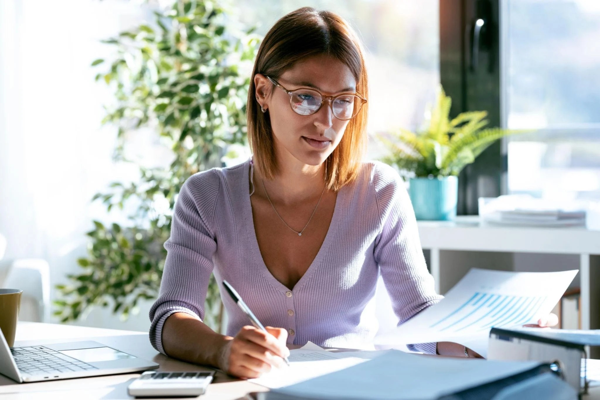 A woman with shoulder-length brown hair and glasses, wearing a light purple cardigan, writing on paper at a desk in a bright room with large windows and green plants in the background.