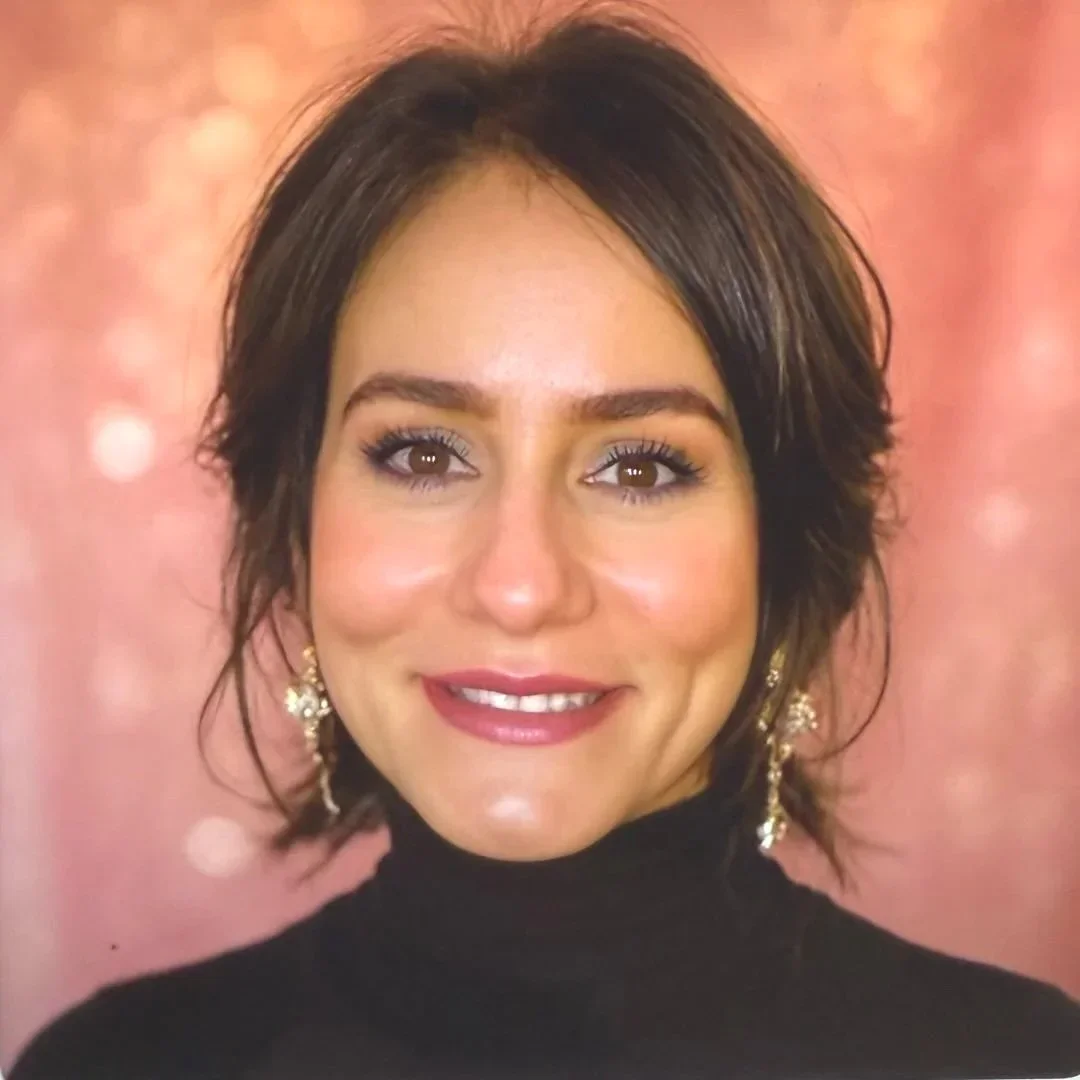 A woman with short dark brown hair in loose waves, wearing makeup, earrings, and a black turtleneck, smiling in front of a pink, bokeh background.