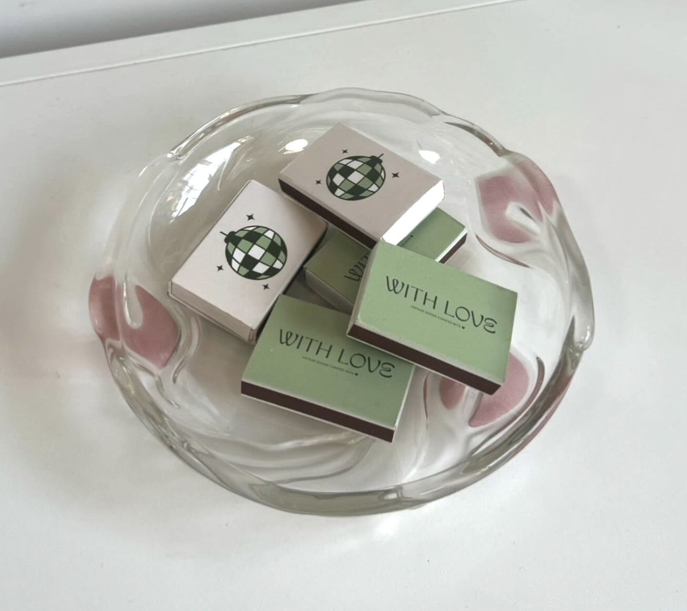 A glass bowl with pink hearts and swirl designs containing matchboxes, some labeled 'WITH LOVE' and others with a globe illustration.
