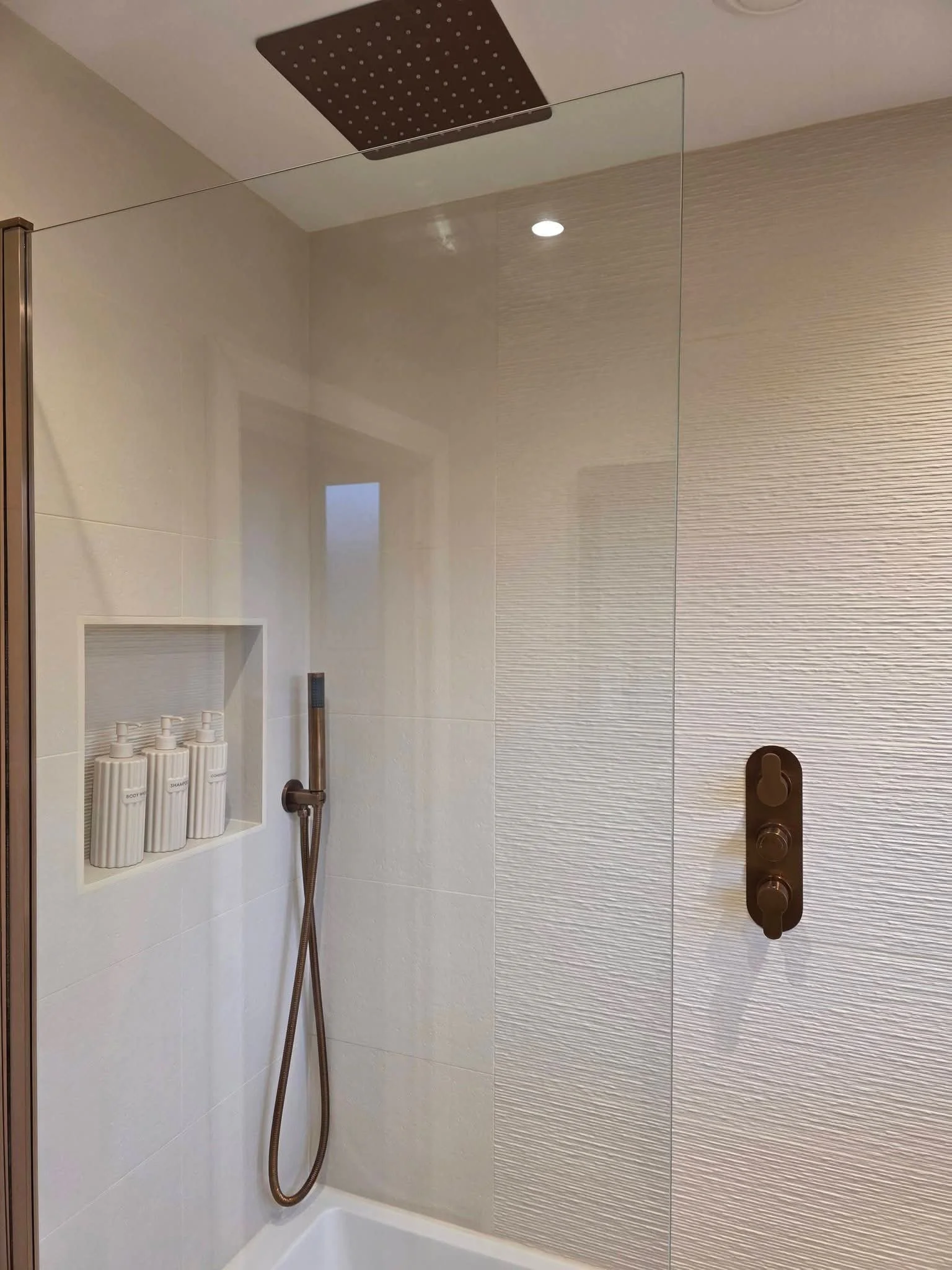Modern shower with a rectangular head, handheld spray, built-in soap and shampoo holders, and a dark bronze control panel with three knobs.