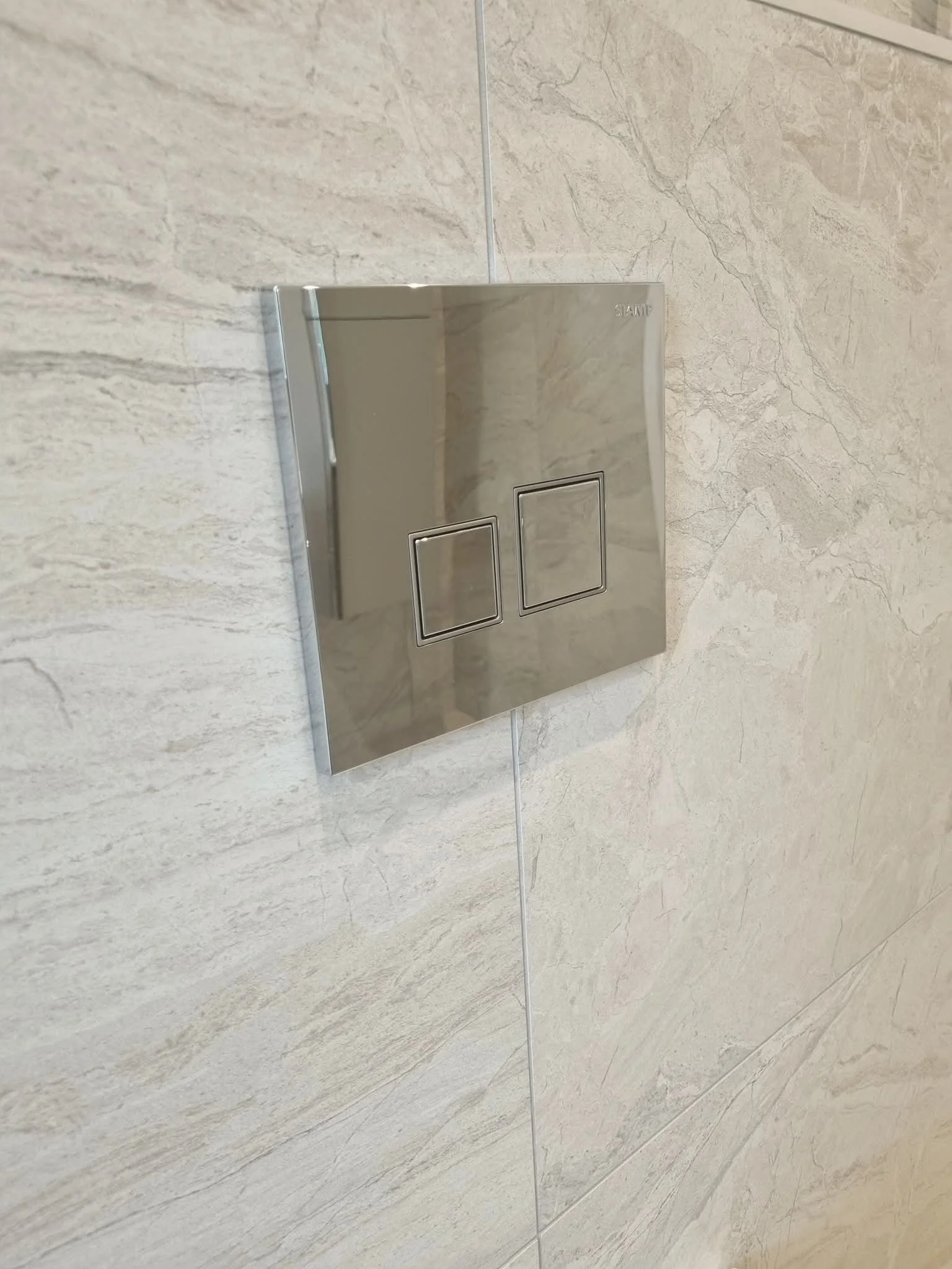 Chrome flush plate fitted into a light stone-effect tiled wall above a contemporary toilet.