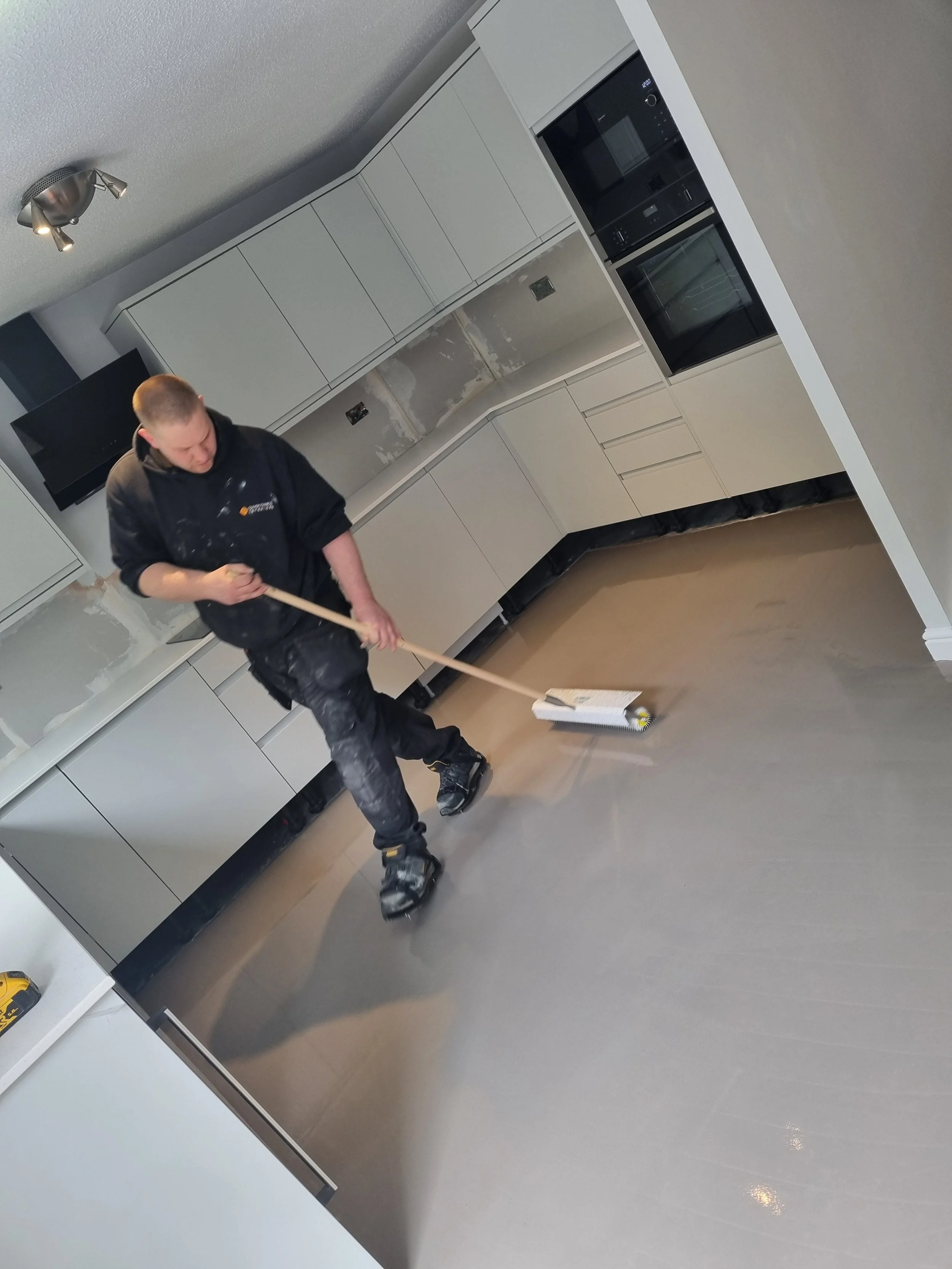 A person is sweeping a kitchen floor with a broom, wearing a black hoodie and black pants with paint stains.