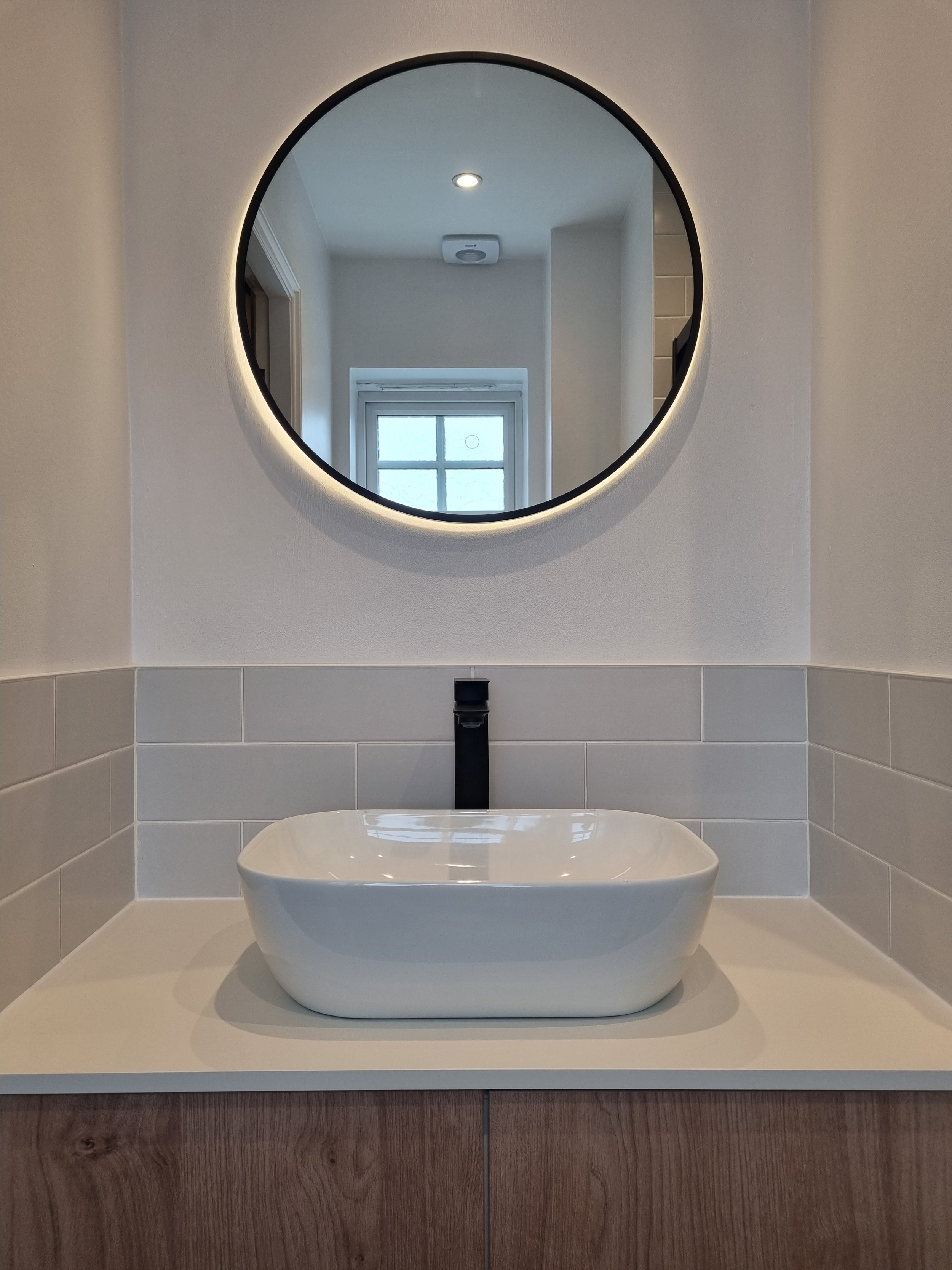 Modern bathroom renovation featuring a round illuminated mirror, white countertop basin and warm wood-effect vanity unit.