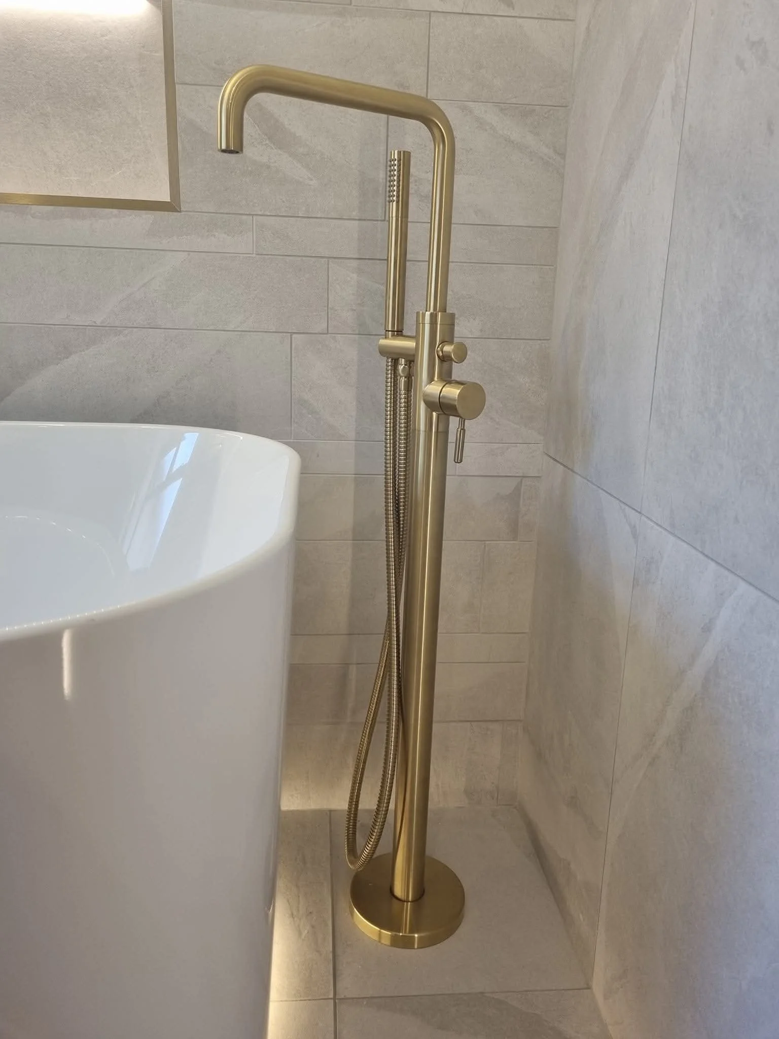 Contemporary freestanding white bathtub with brushed gold floor-mounted tap and neutral stone-effect wall tiles.