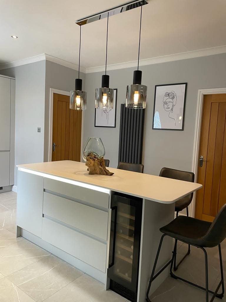 Modern kitchen island with a light wooden countertop, a built-in wine fridge, and brown chairs. Overhead pendant lights with glass covers hang above the island. The background features two wooden doors, black wall art, and artwork of line drawings on the wall.