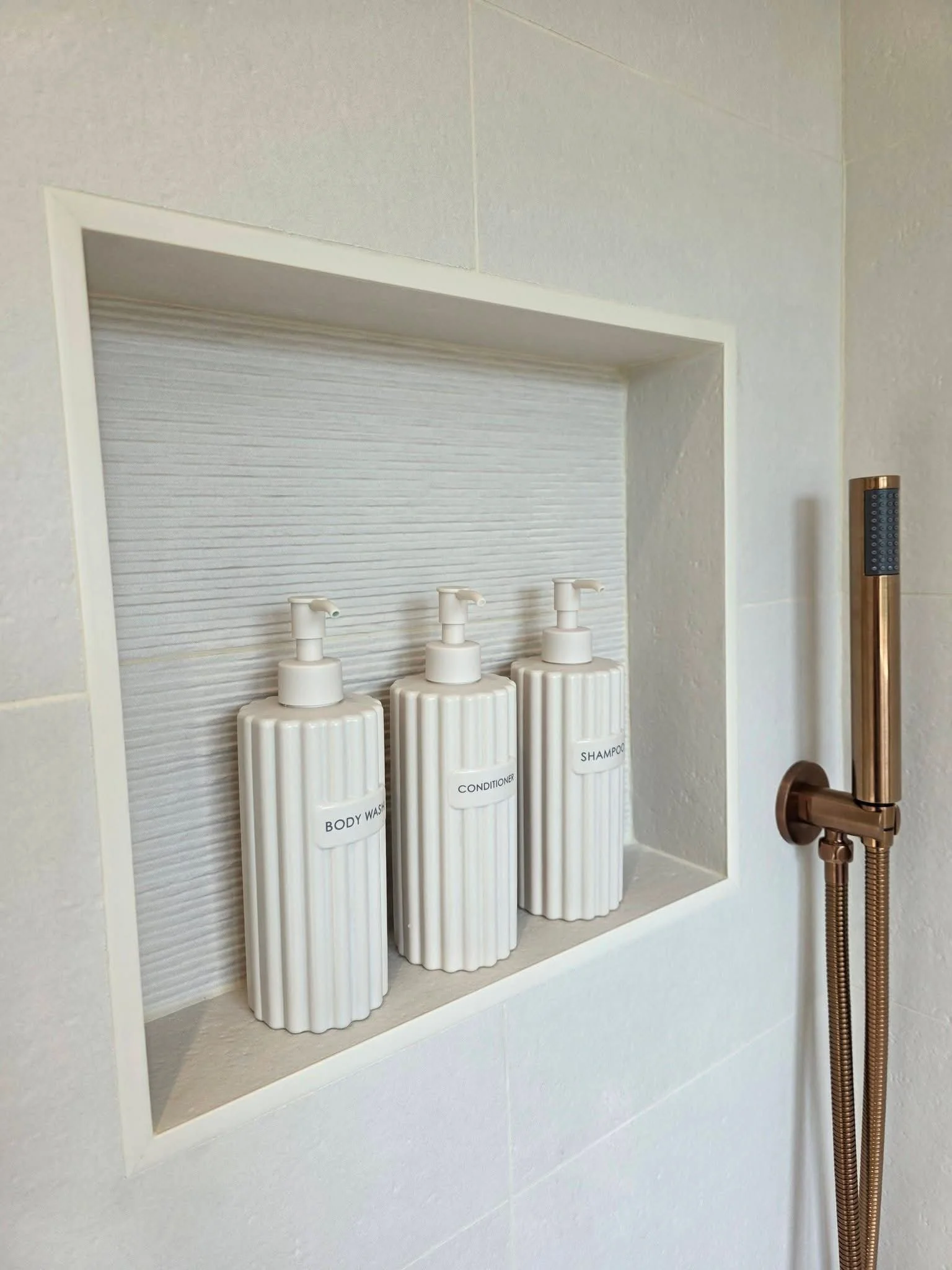 Built-in shower niche with white toiletry bottles, textured wall tiles and brushed bronze shower fittings.