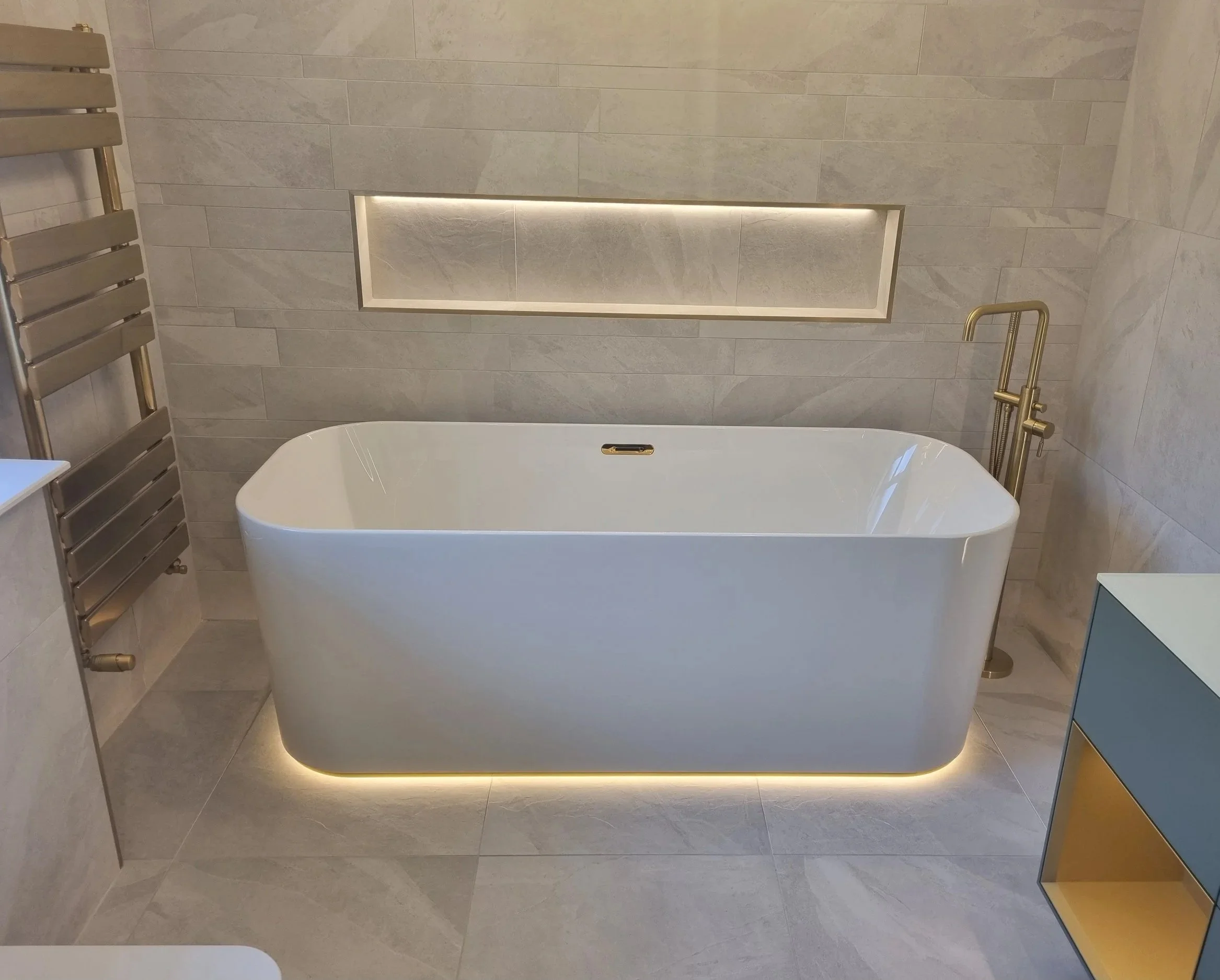 Modern bathroom renovation by Oliver Francis Interiors featuring a freestanding bath, grey stone-effect tiles, recessed lighting and brushed gold fittings.