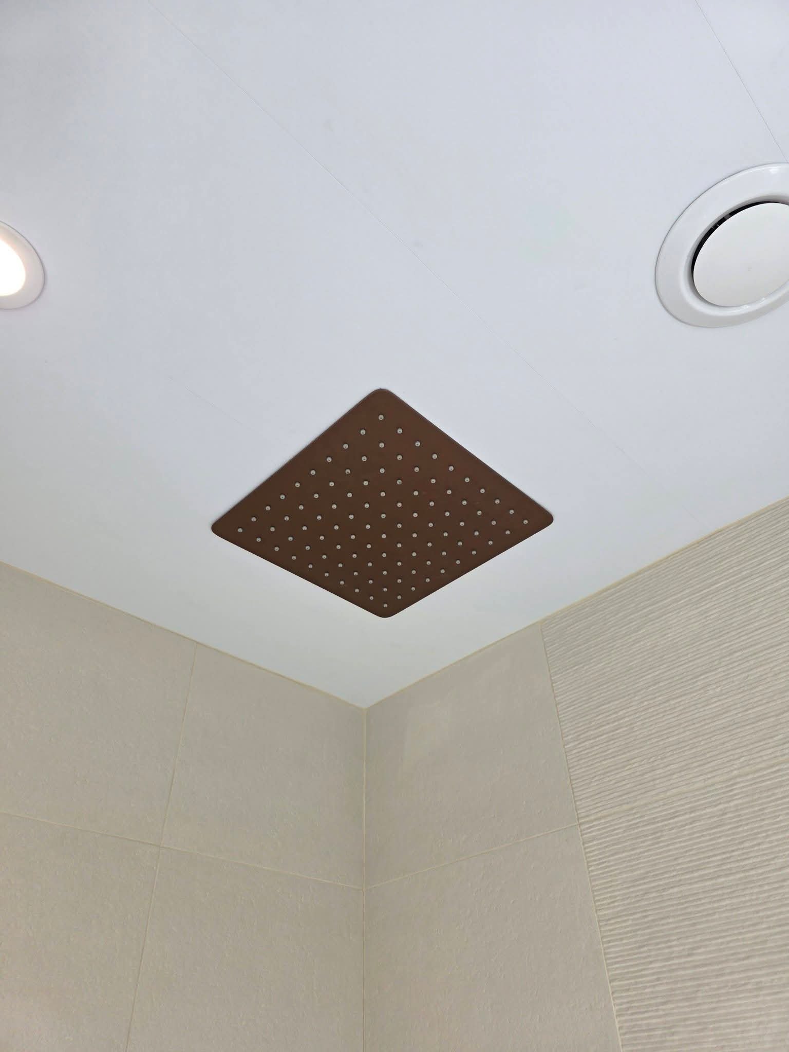 Square brushed bronze rainfall showerhead mounted in the ceiling above a contemporary tiled shower.