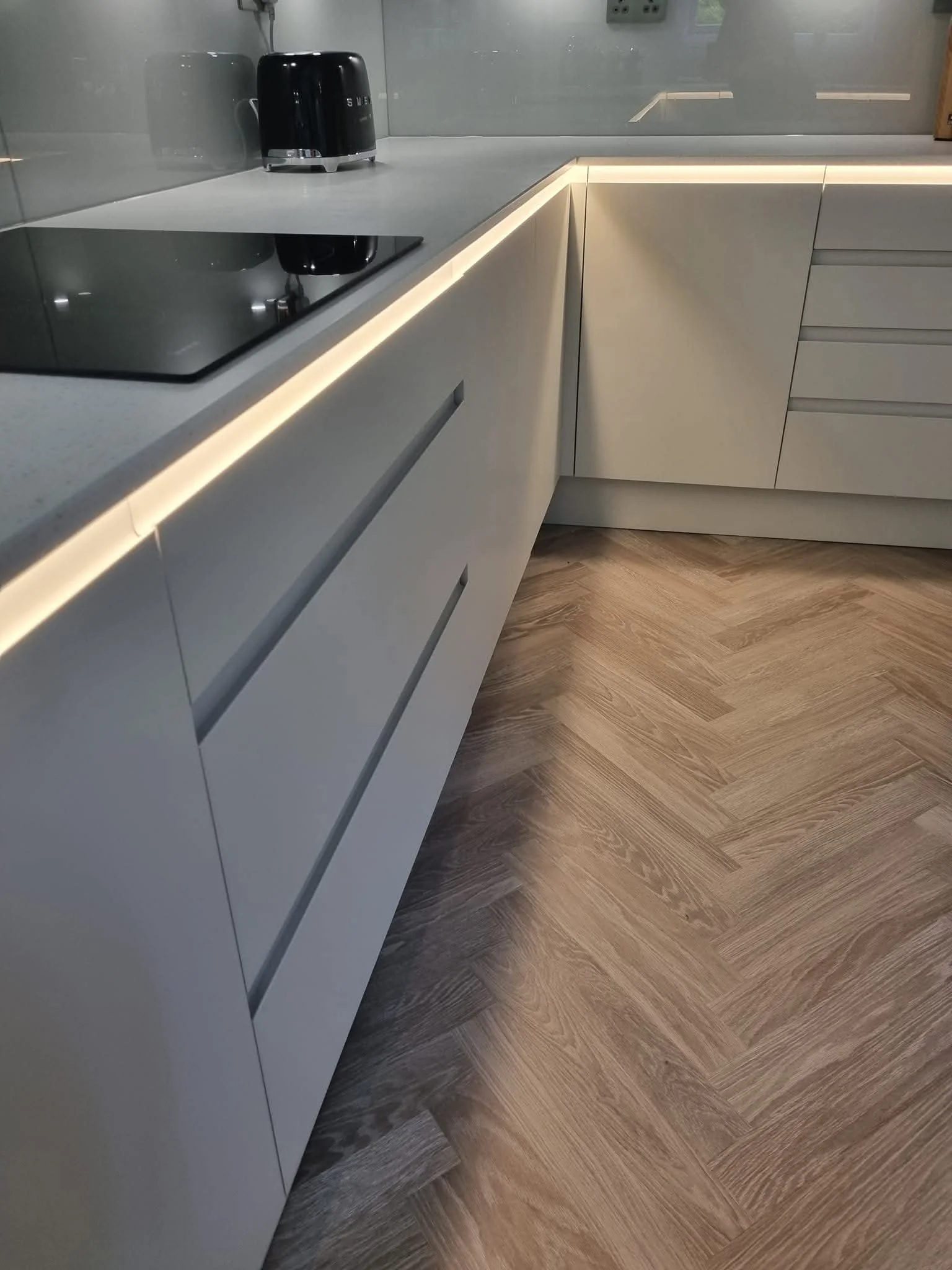 Close-up of modern handleless kitchen cabinetry with sleek worktops, glass splashbacks and warm LED lighting.